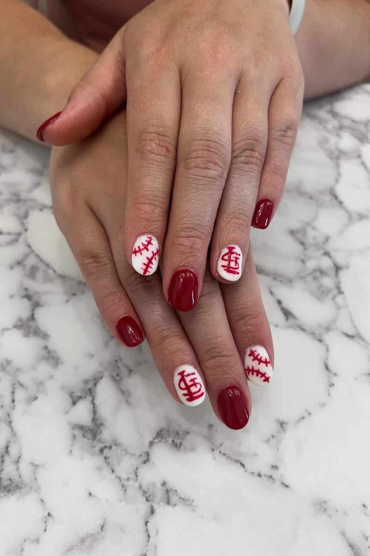 Hands with red and white painted nails, some featuring baseball stitching and a red logo resembling the St. Louis Cardinals emblem, resting on a marble surface.