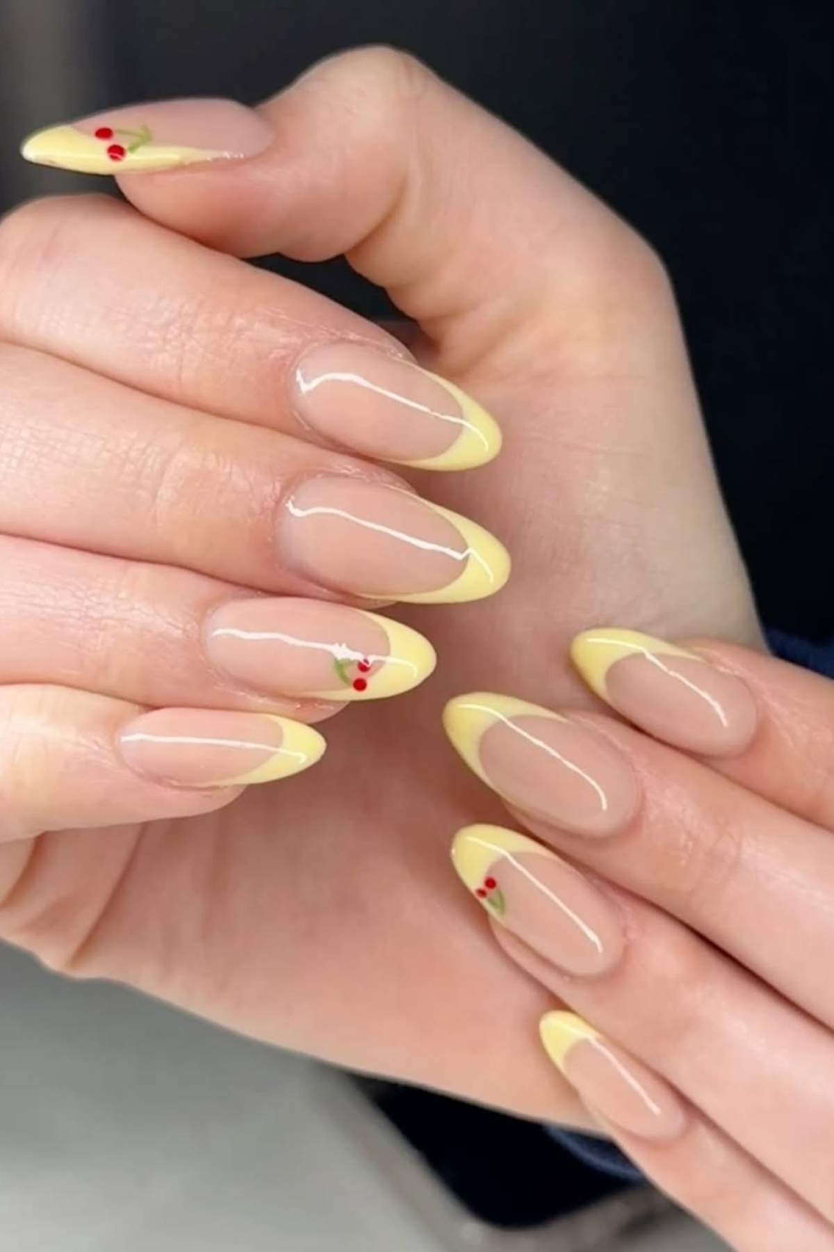 A close-up of hands with almond-shaped nails featuring light yellow French tips and small red cherry designs on two accent nails.