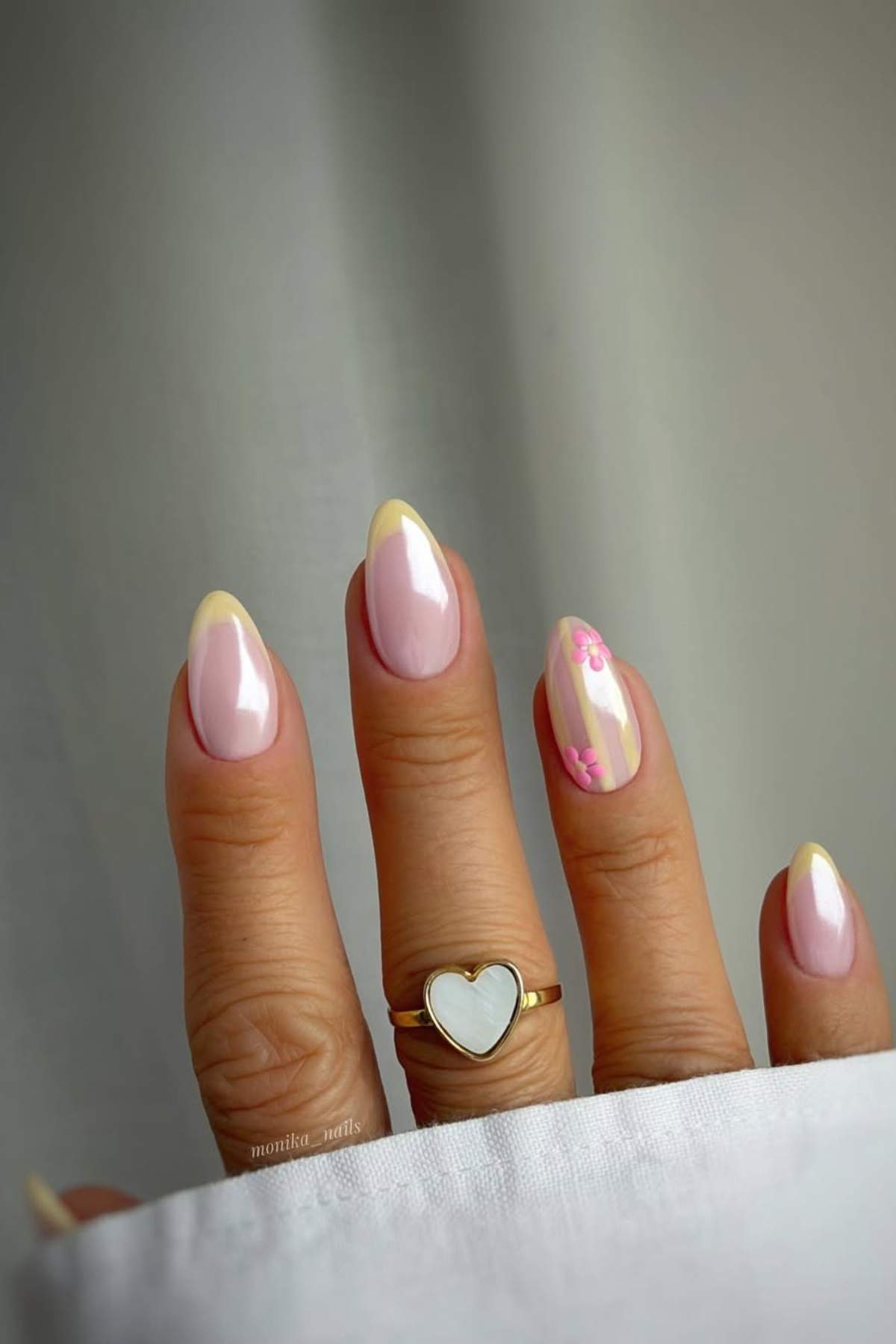 A hand with almond-shaped nails painted in pale pink with yellow French tips; one nail has pink floral designs. The person wears a gold ring with a white heart.