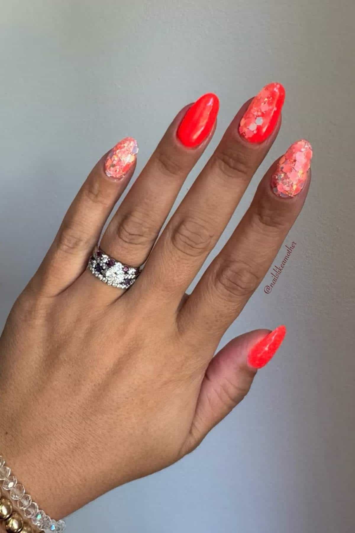 A hand with long, almond-shaped nails painted bright red and glittery pink, wearing a diamond ring and gold bracelets, resting against a light background.