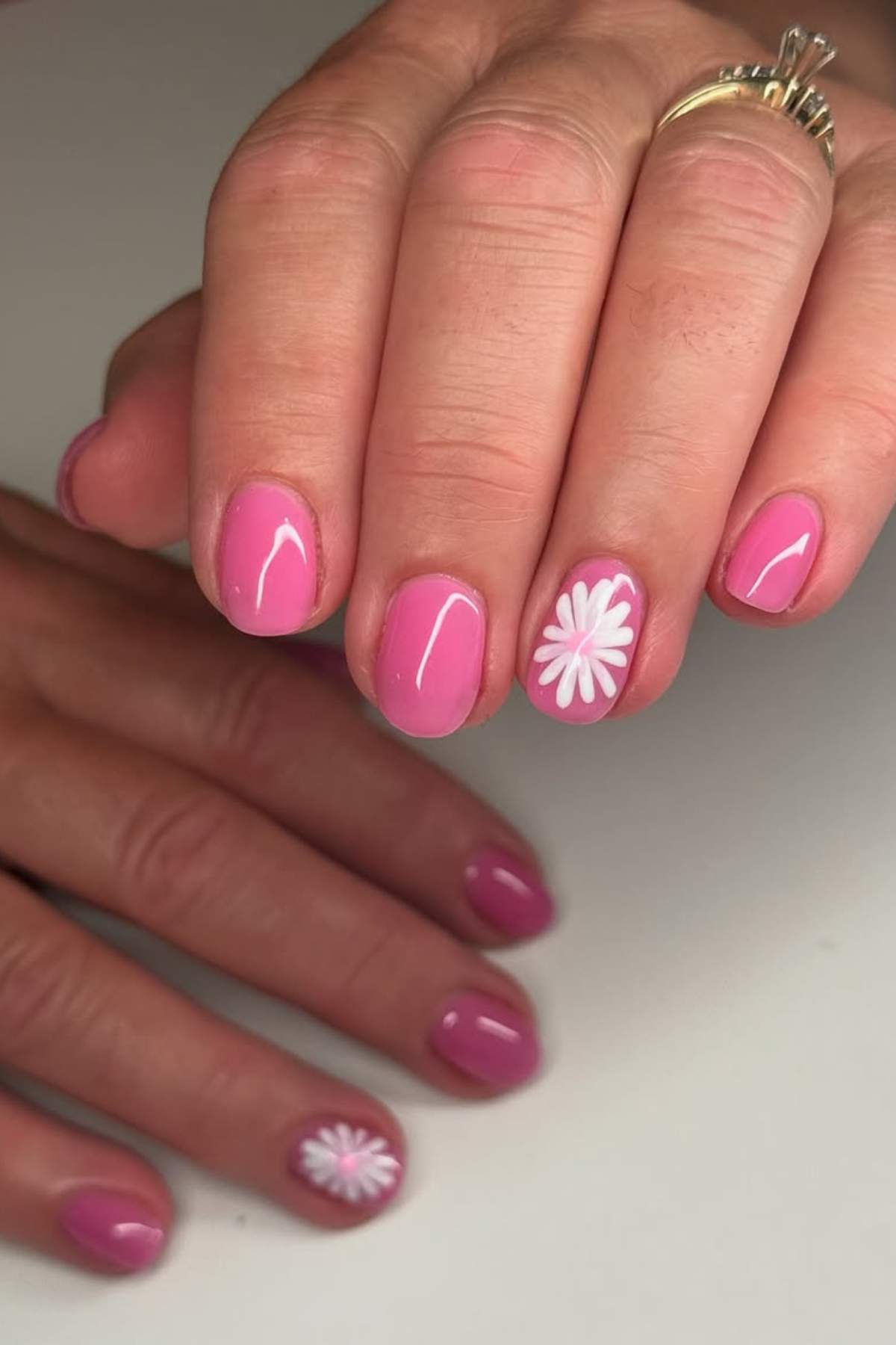 A hand with short, oval-shaped pink nails; one nail on each hand features a white and yellow daisy design.