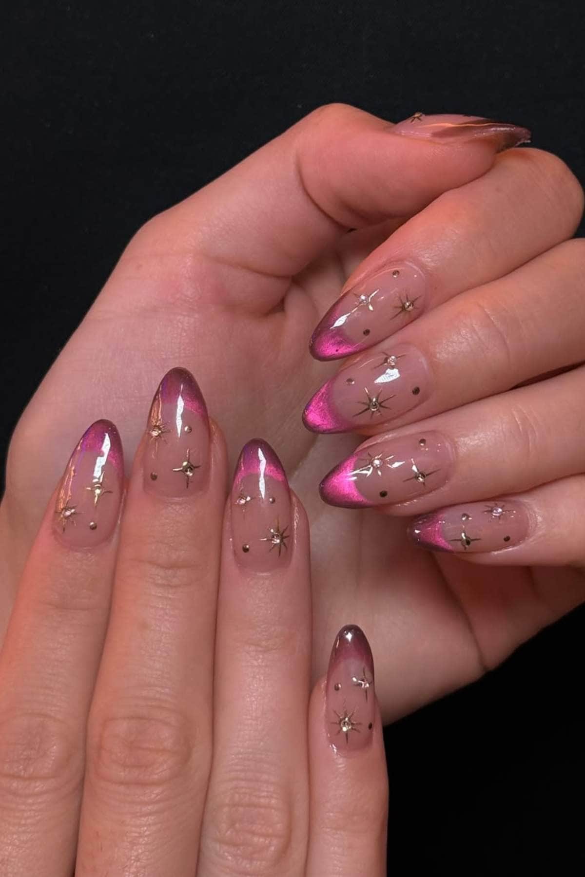 Hands with almond-shaped nails featuring a pink gradient, gold star accents, and small black dots on a clear base, set against a black background.