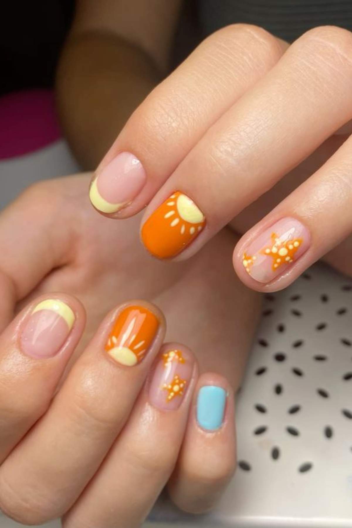 Close-up of hands with manicured nails featuring pastel yellow French tips, orange nails with sun designs, star patterns, and one nail painted solid light blue.