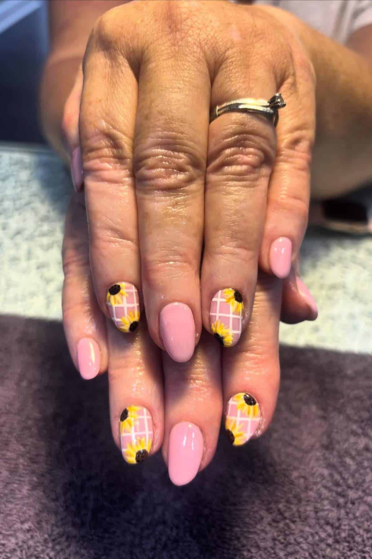 A hand with pink manicured nails; some nails feature a grid pattern with sunflower designs on a light background.