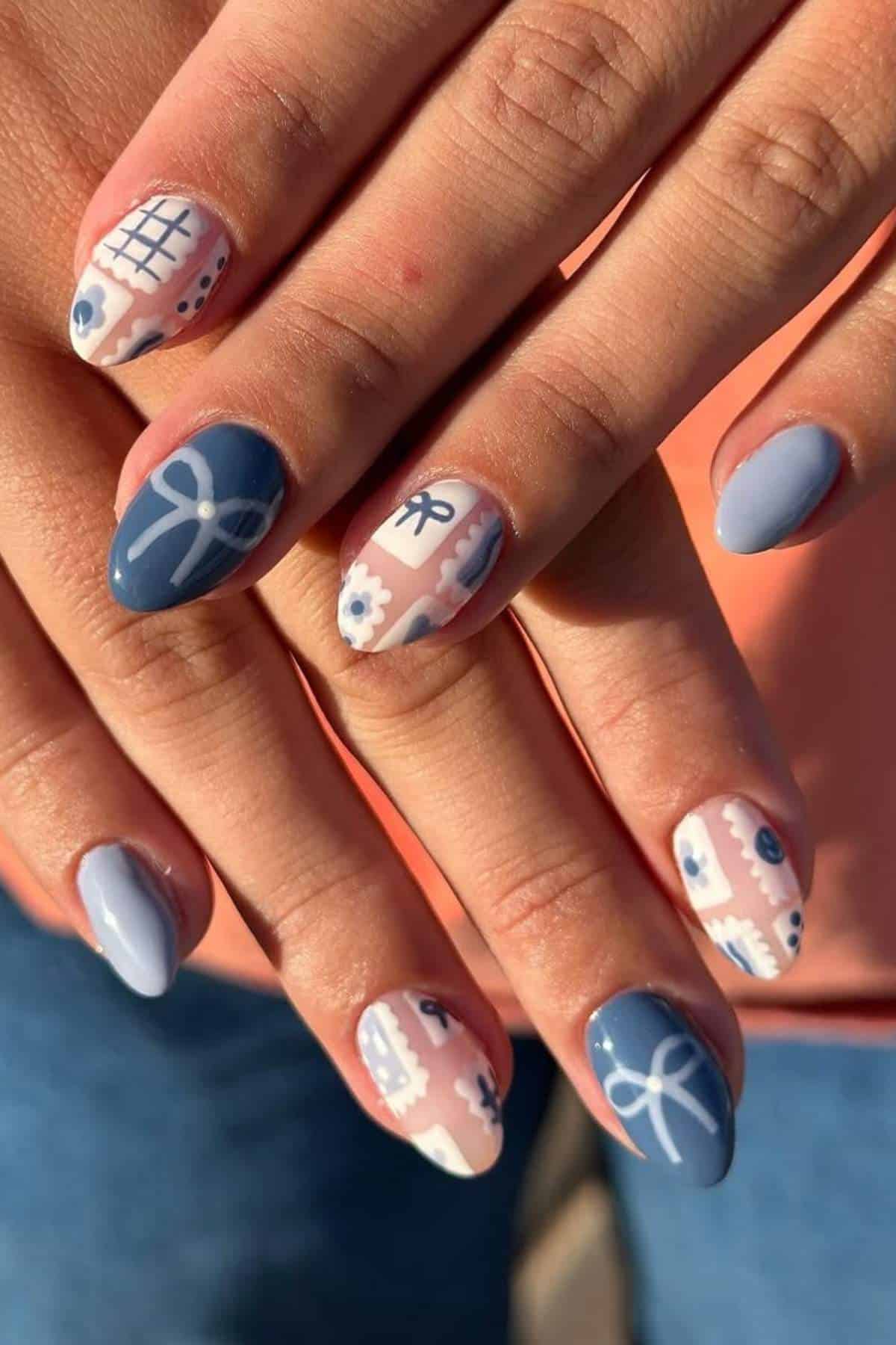 Close-up of hands with almond-shaped nails, painted in light blue and white with floral, grid, and ribbon patterns in a mix of solid and intricate designs.
