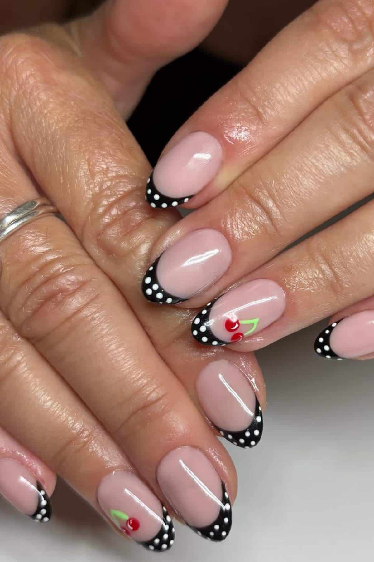 Hands with almond-shaped nails featuring black French tips with white polka dots and small cherry designs on two nails.