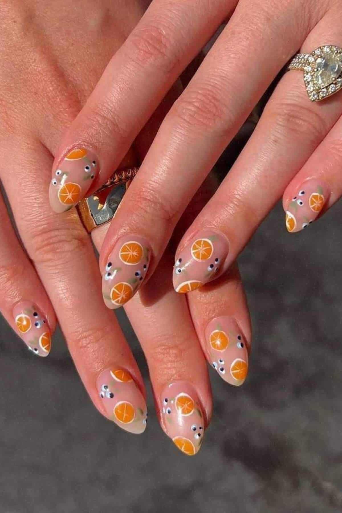 A person's hands with almond-shaped nails featuring orange slice and black dot nail art on a neutral background.