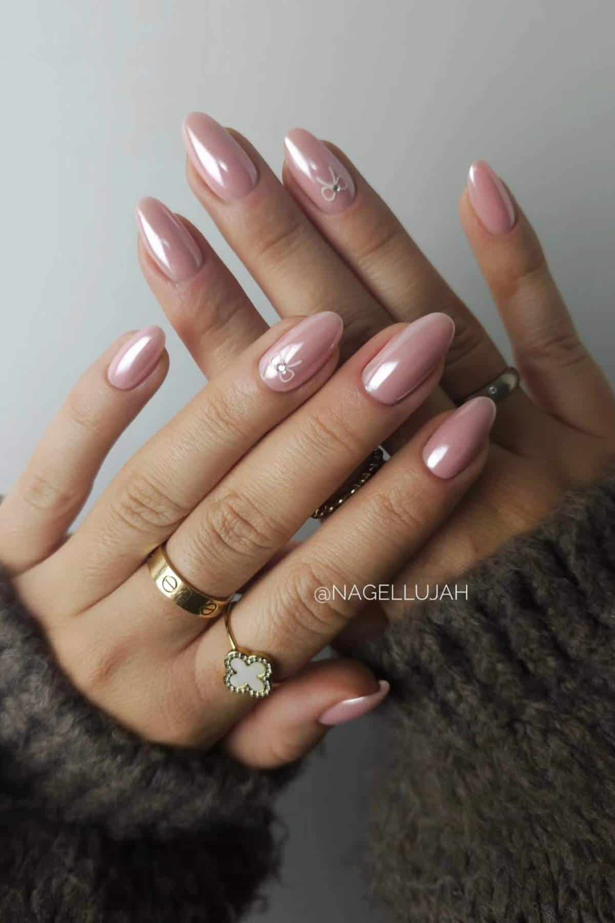 Hands with almond-shaped, glossy light pink nails; two nails feature small white bow designs. The person wears gold rings and a brown textured sweater.