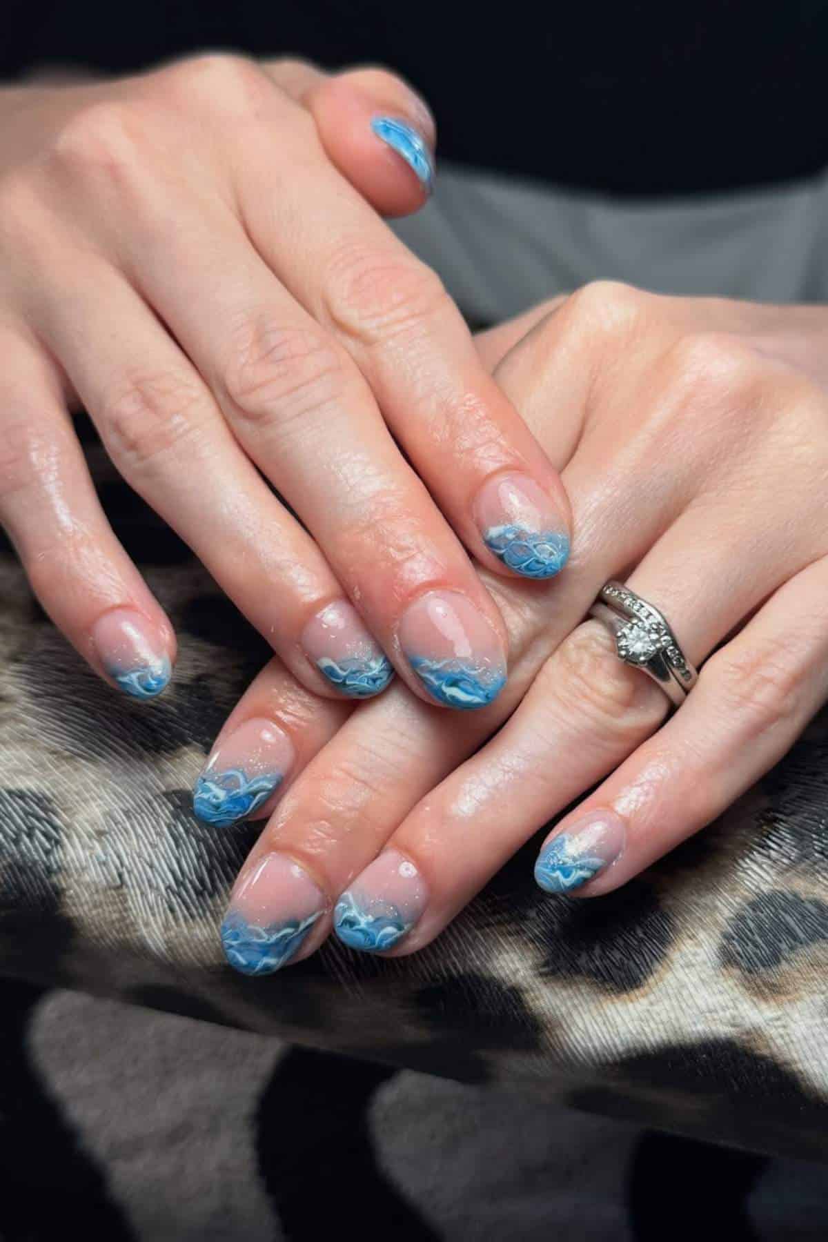 Hands with short, rounded nails featuring blue and white wave nail art on the tips. One hand wears a silver ring with a clear stone. The background is a leopard print fabric.
