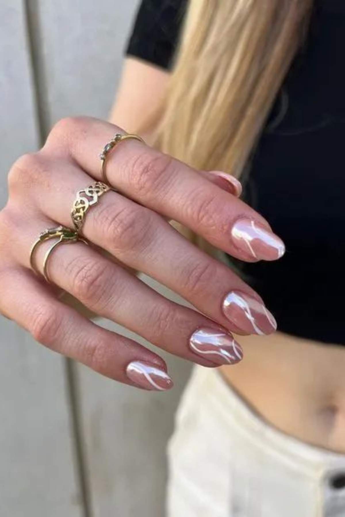 A hand with long, almond-shaped nails painted with a glossy, light pink polish and white abstract lines, wearing several gold rings, is shown against a neutral background.