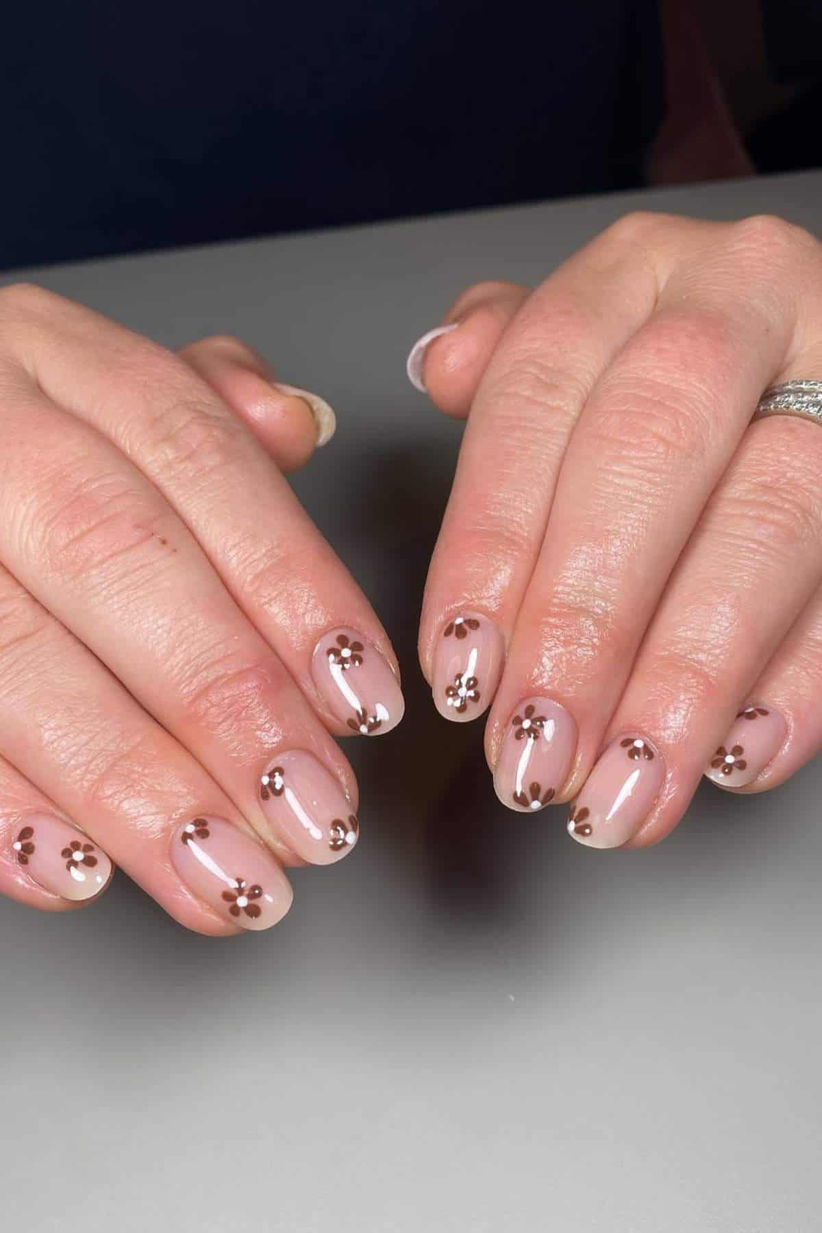 Hands with short, oval-shaped nails featuring a nude base and small brown and white daisy flower nail art on each nail.