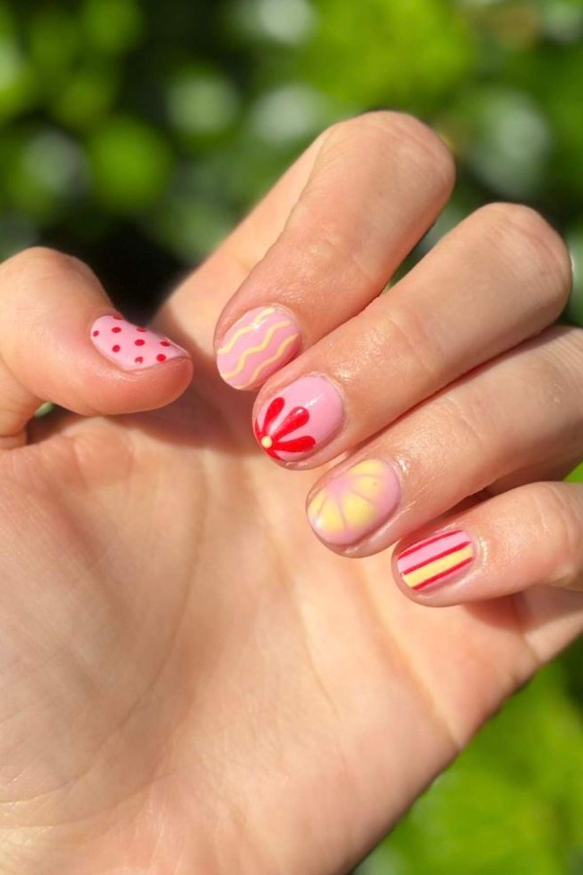 A hand with pastel pink nail polish, each nail featuring a different red and yellow abstract or geometric design, shown against a green blurred background.