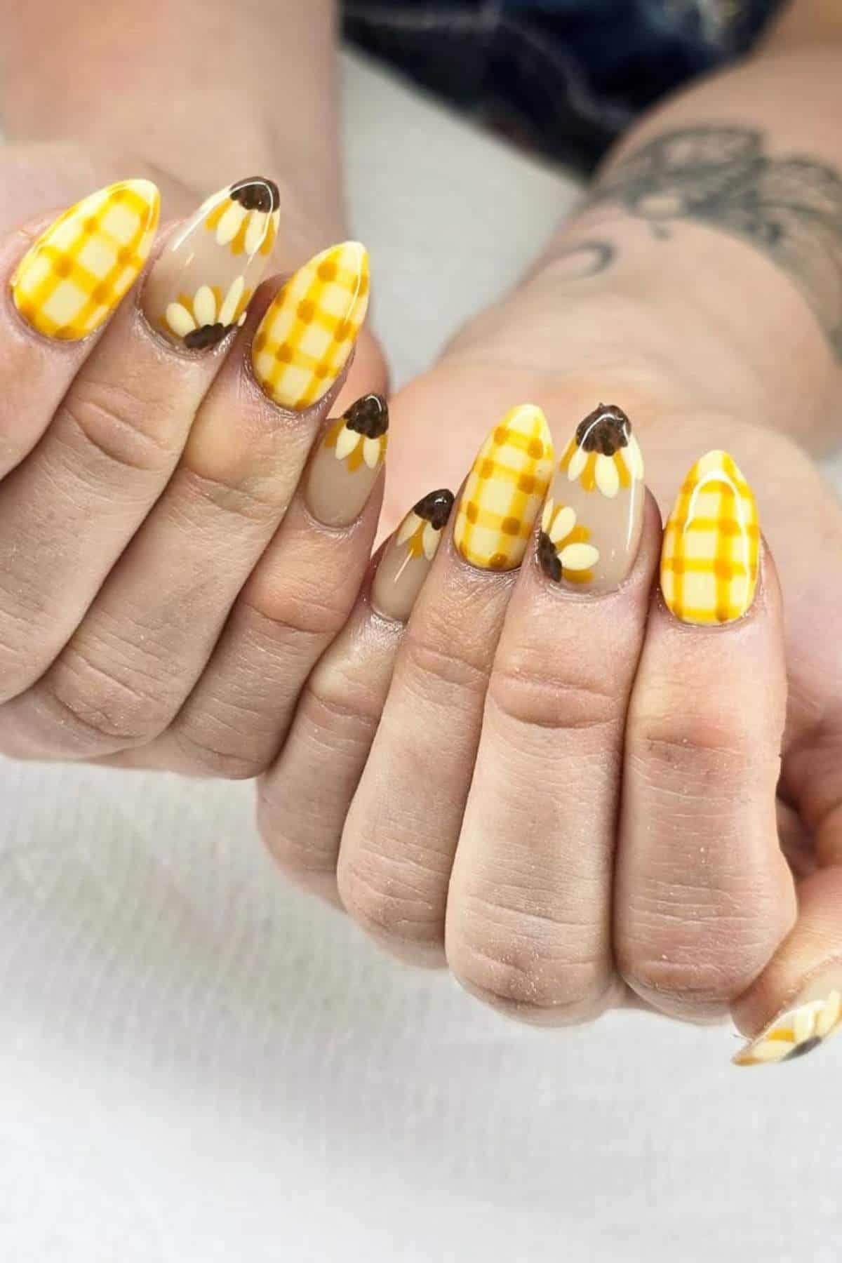 Hands with long almond-shaped nails featuring yellow gingham patterns and daisy flower designs on a light background.