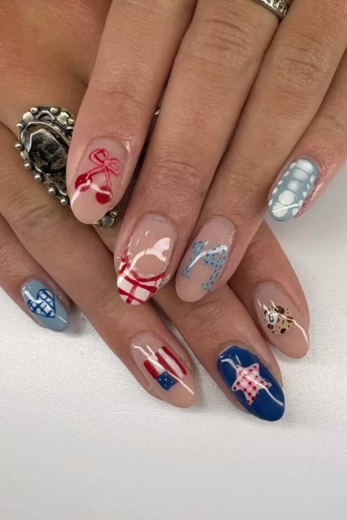 A hand with manicured nails featuring various designs, including cherries, hearts, stars, a flag, checks, polka dots, and a cookie, in red, blue, white, and beige tones.