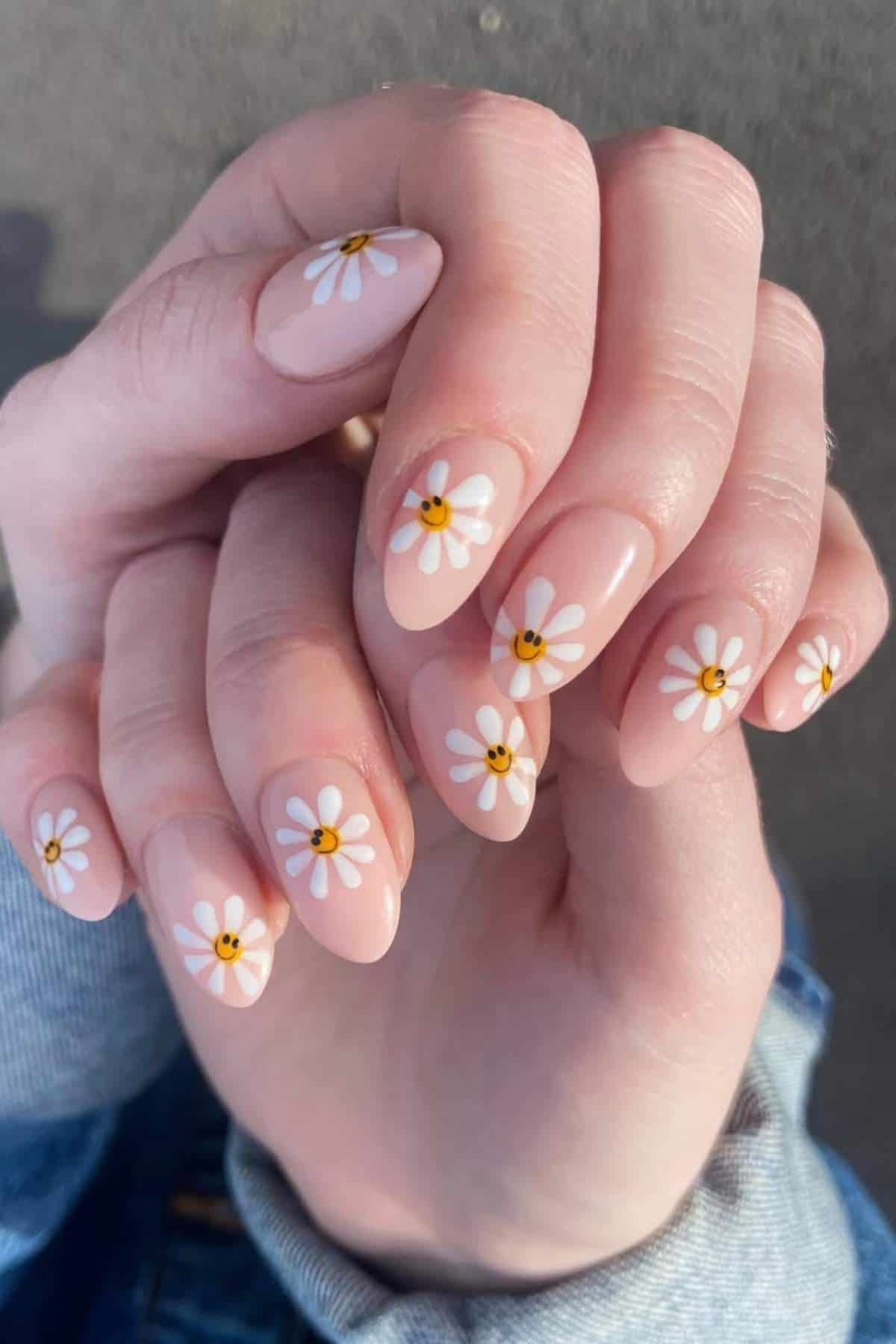 Close-up of hands showing fingernails painted with a light pink base and decorated with white daisy flower designs with yellow centers.