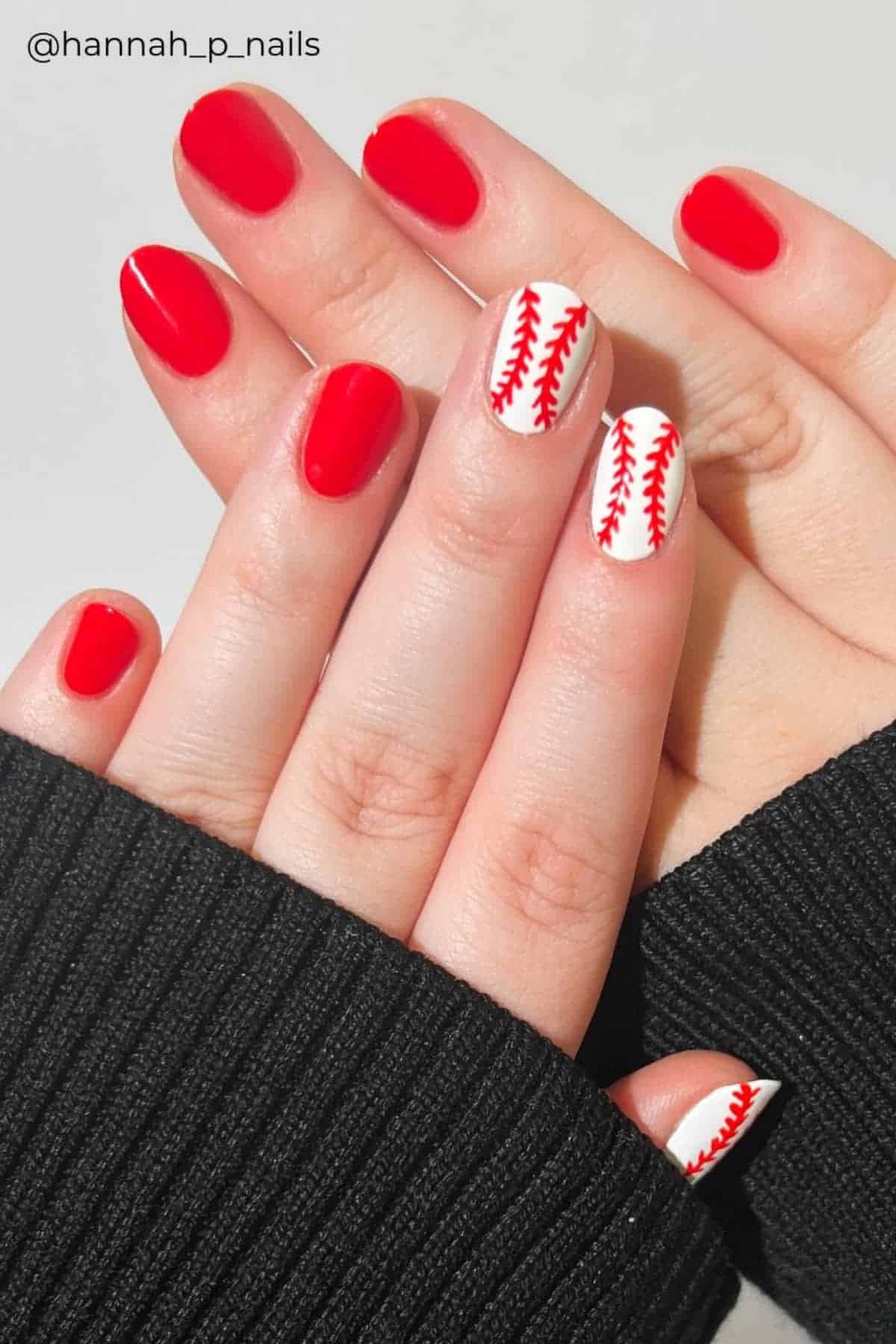 Hands with red nail polish and two accent nails featuring white polish with red baseball stitching designs.