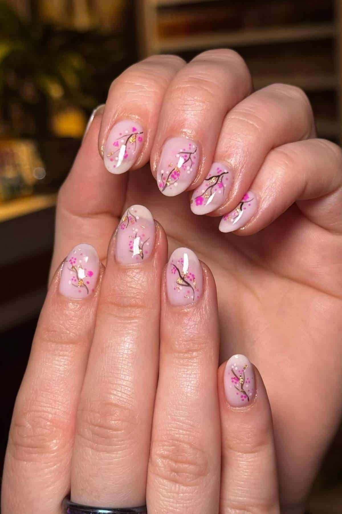A close-up of hands with almond-shaped nails featuring pink and gold floral nail art on a translucent nude base.