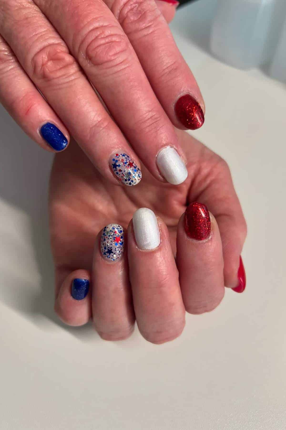 A hand with a patriotic manicure featuring red, white, and blue nails with glitter and star accents.