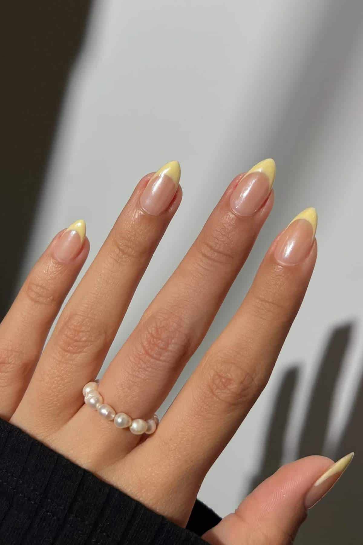 A hand with almond-shaped nails painted with a pale yellow French tip manicure, wearing a pearl beaded ring, against a neutral background.