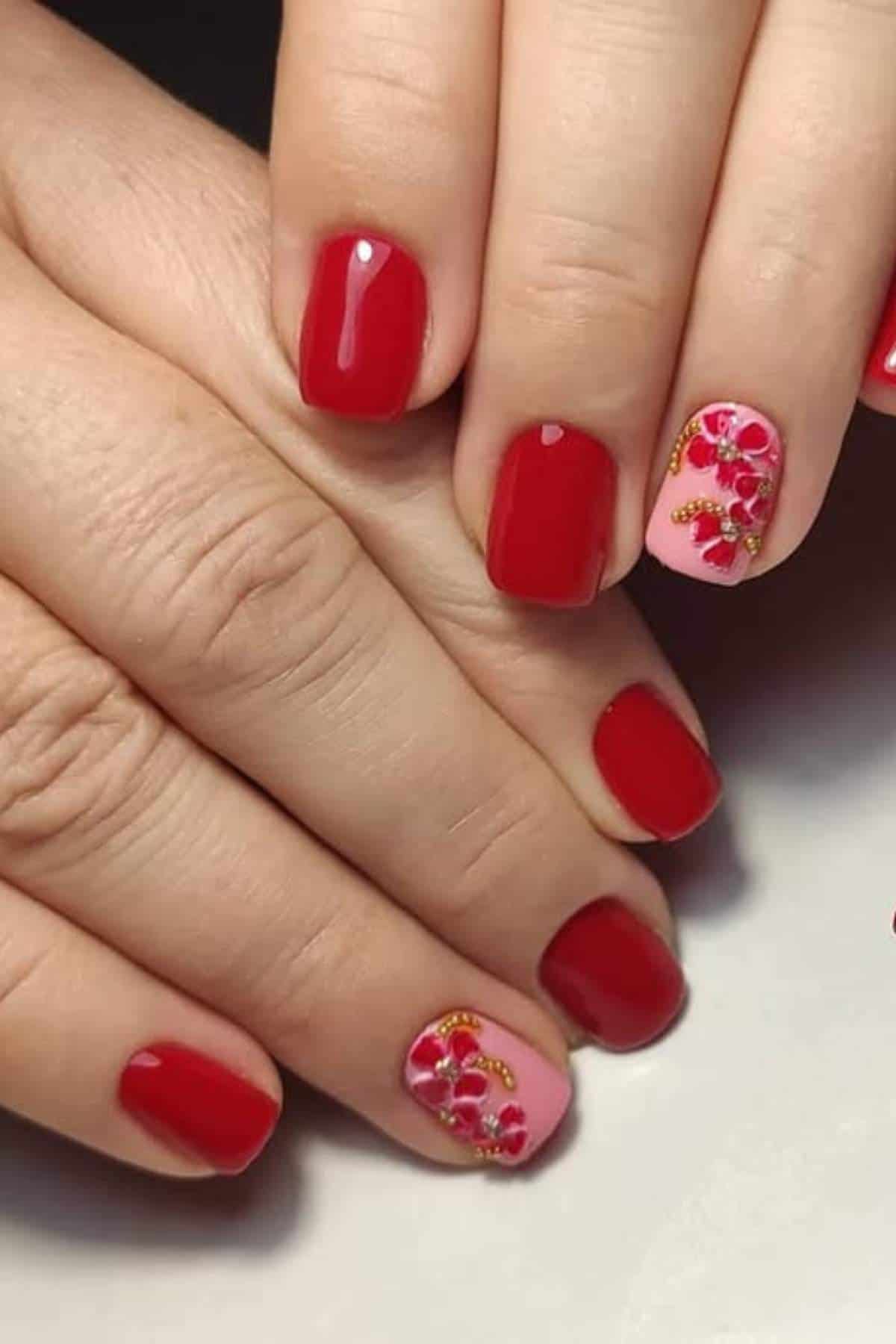 Hands with short, square-shaped red nails; one nail on each hand is painted light pink and decorated with intricate floral designs and small embellishments.