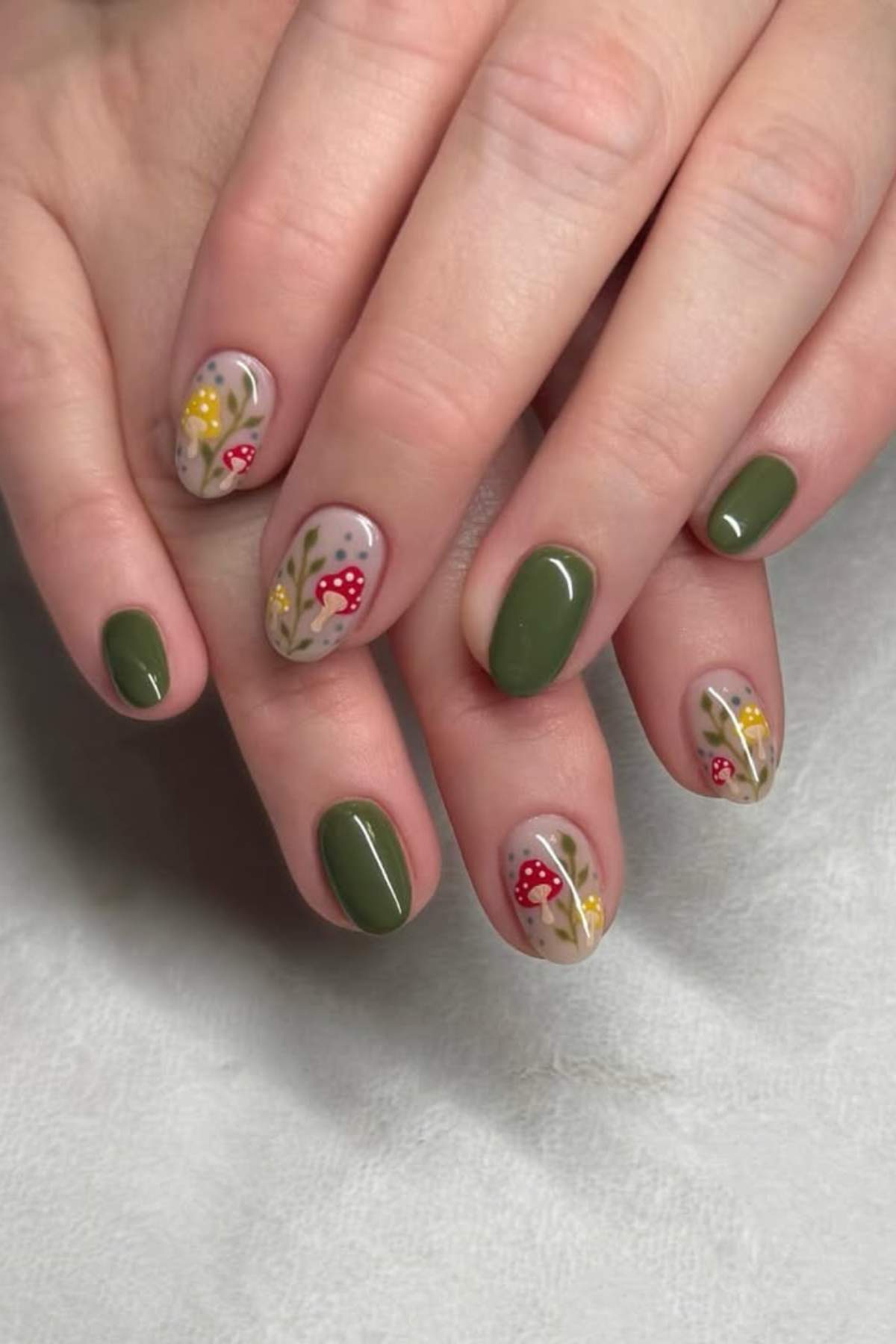 Hands with short, oval-shaped nails painted olive green, with accent nails featuring red and yellow mushroom and floral nail art on a nude base.