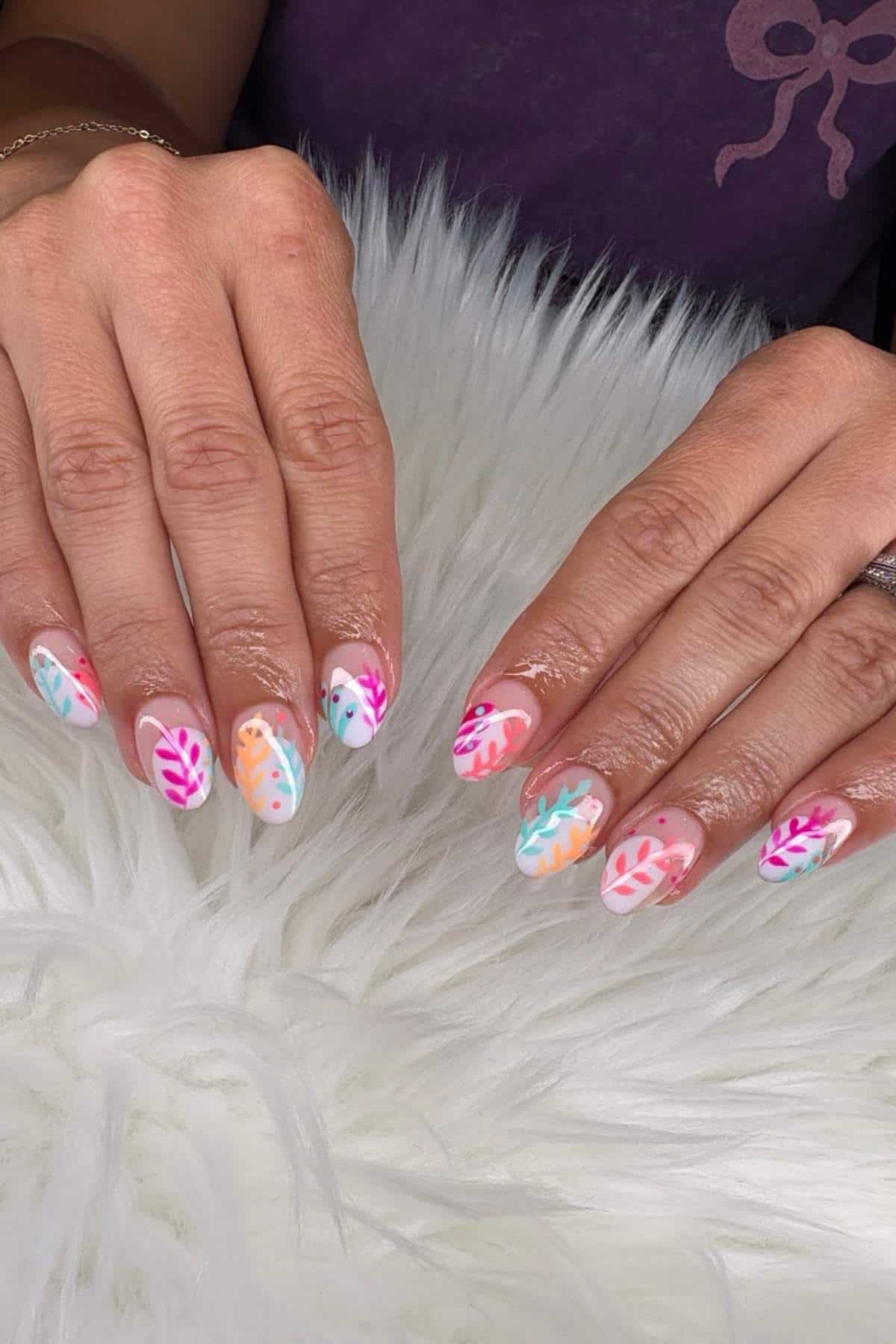 Hands with almond-shaped nails featuring colorful leaf designs in pink, blue, orange, and purple on a white background, displayed on a white furry surface.