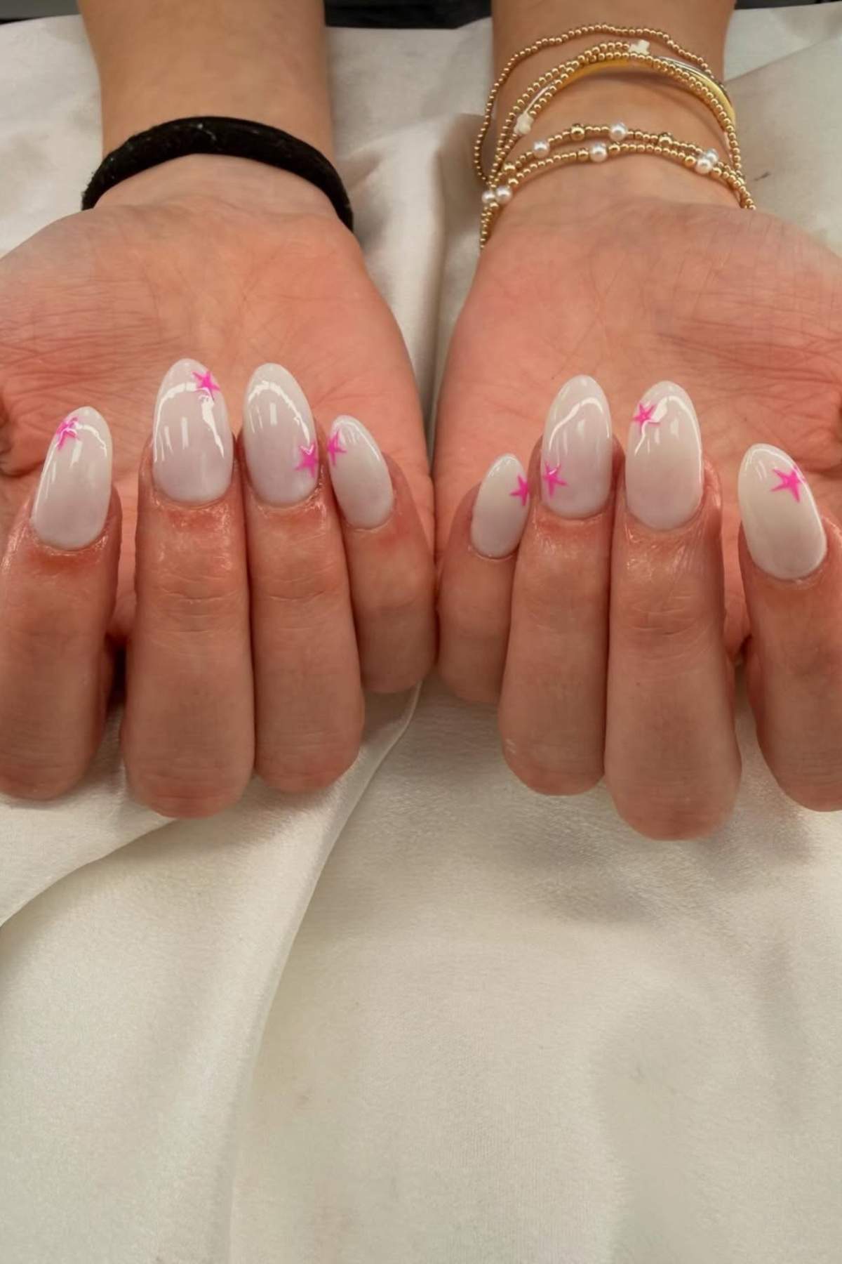 Two hands with almond-shaped, pale pink nails featuring small pink star designs on each nail. The person is wearing gold and beaded bracelets on one wrist.