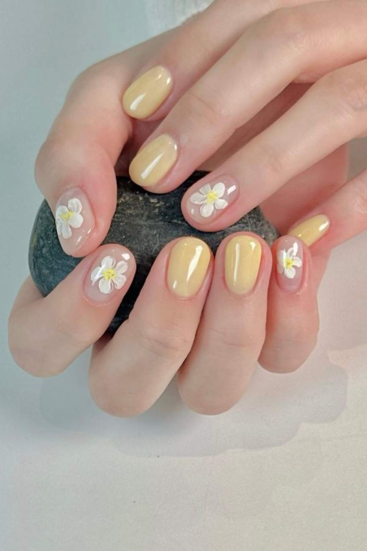 Hands with short yellow nails, some featuring white daisy flower nail art, holding a smooth black stone.