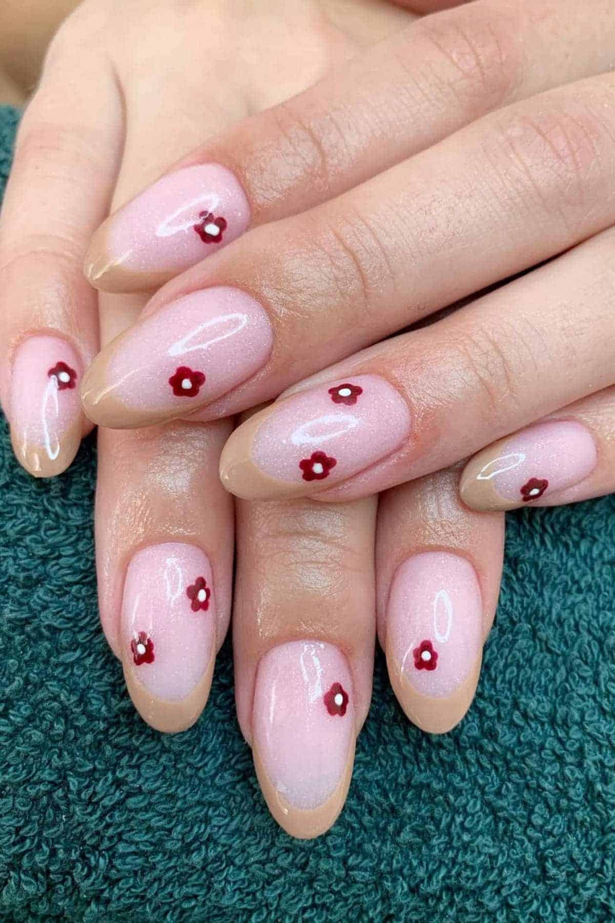 Hands with almond-shaped nails painted light pink, each nail featuring a small red flower design, resting on a green textured fabric.