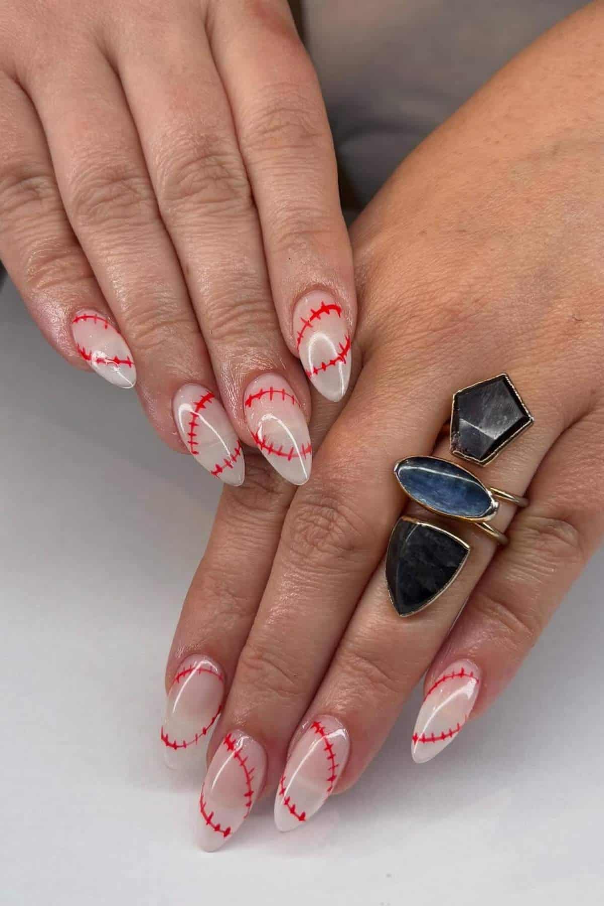 Hands with almond-shaped nails featuring a baseball stitch design in red on a nude base. One hand wears three statement rings with dark and blue stones.