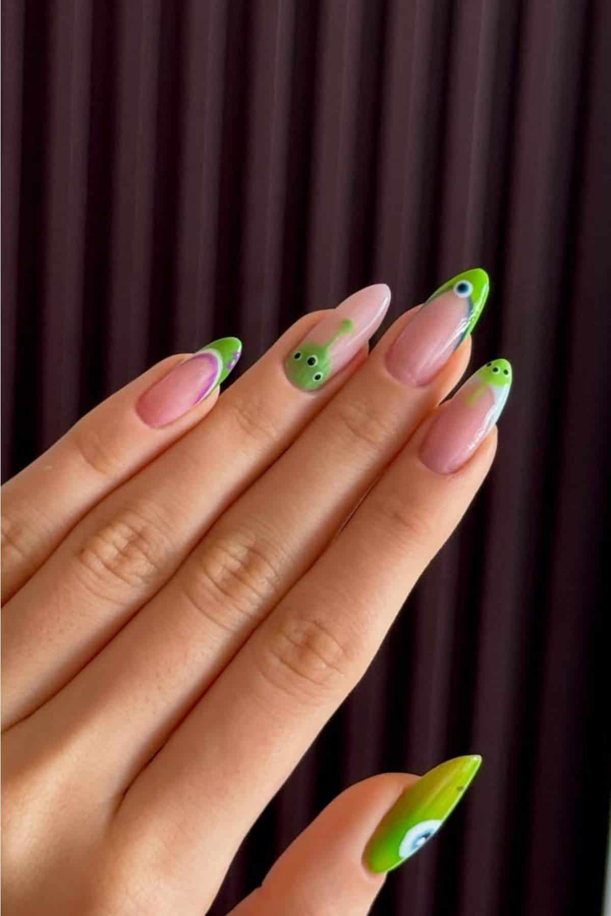 A hand with long almond-shaped nails featuring green and nude nail art, including designs of cute frog faces and wavy patterns.