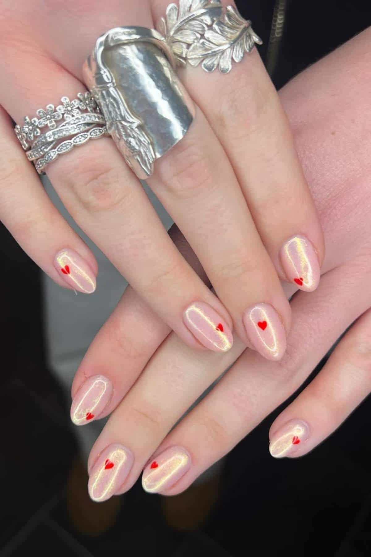 Hands with iridescent nude nail polish and small red heart designs on each nail, wearing multiple silver rings with various textures and patterns.