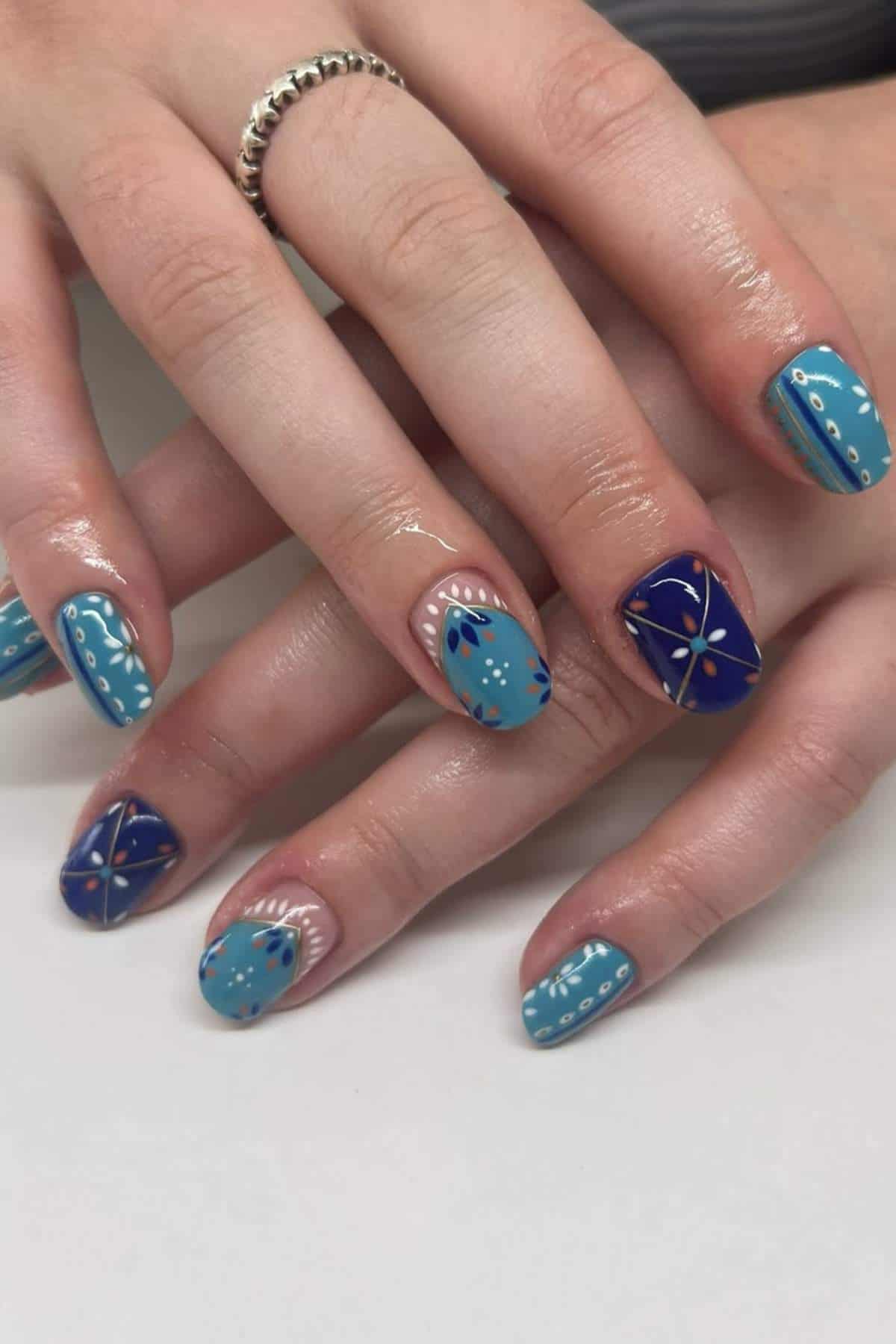 Hands with blue and navy patterned nail art featuring white dots, lines, and floral designs on short, square-shaped nails.