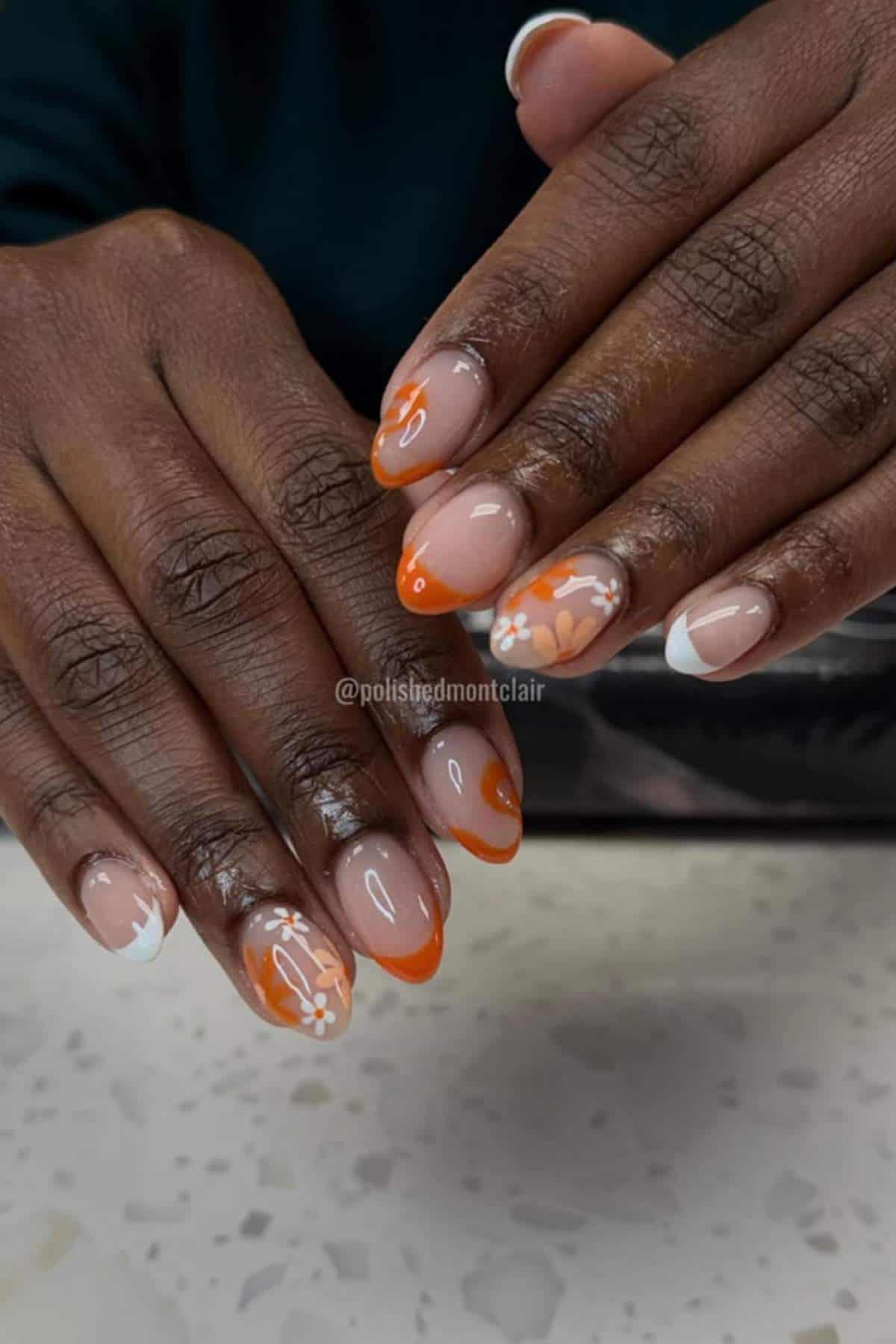 Close-up of hands with almond-shaped nails featuring orange and white floral nail art, French tips, and a nude base.