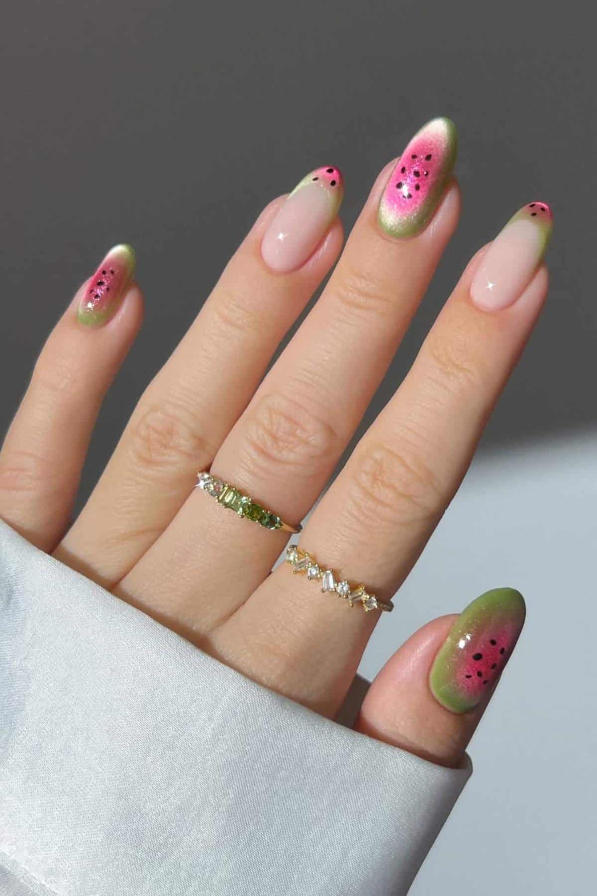 A hand with long, almond-shaped nails painted with a watermelon-inspired design, wearing two rings, against a neutral background.
