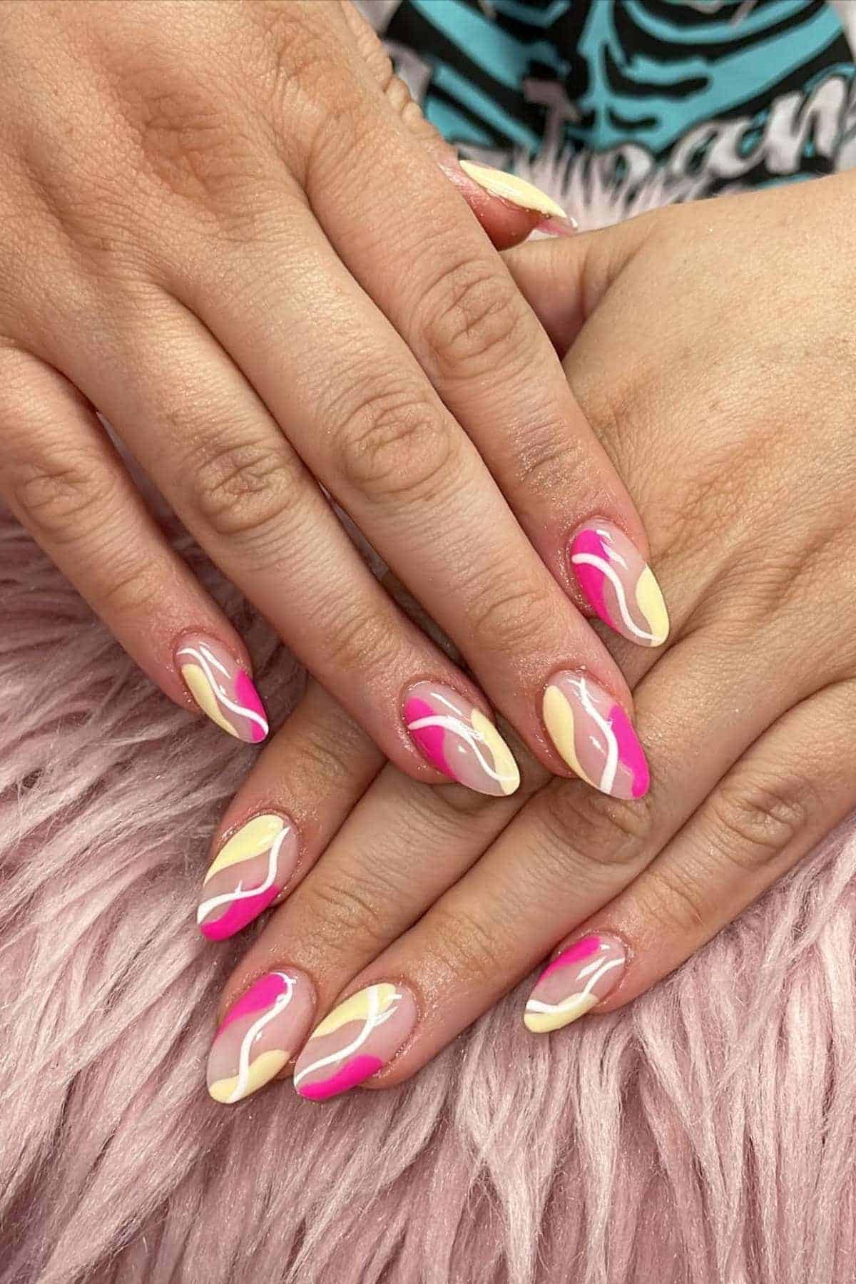 Hands with almond-shaped nails featuring a pink, cream, and white swirl nail art design, resting on a pink fluffy surface.