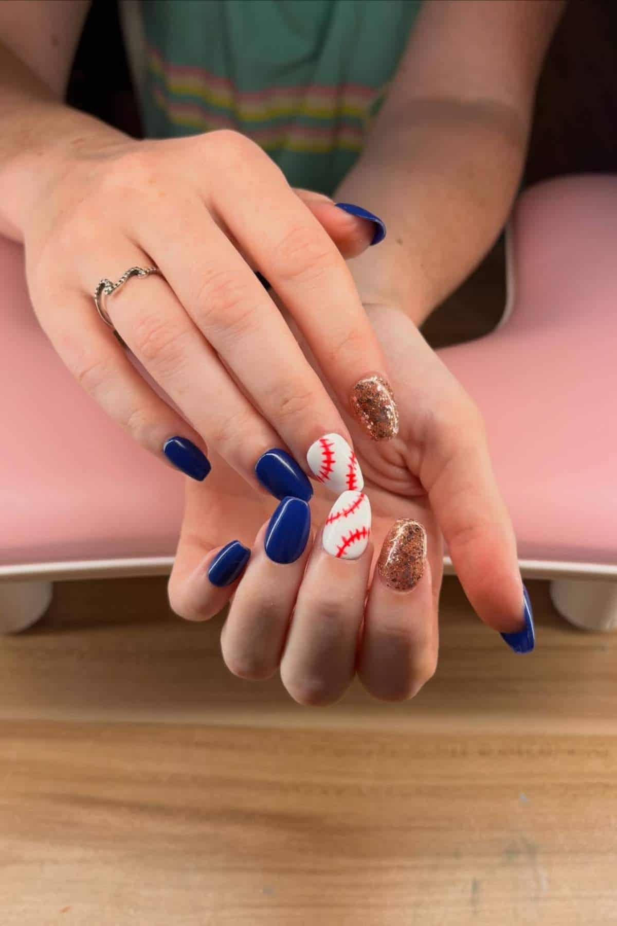 A person displays manicured nails with blue, gold glitter, and white nails featuring red baseball stitching designs.