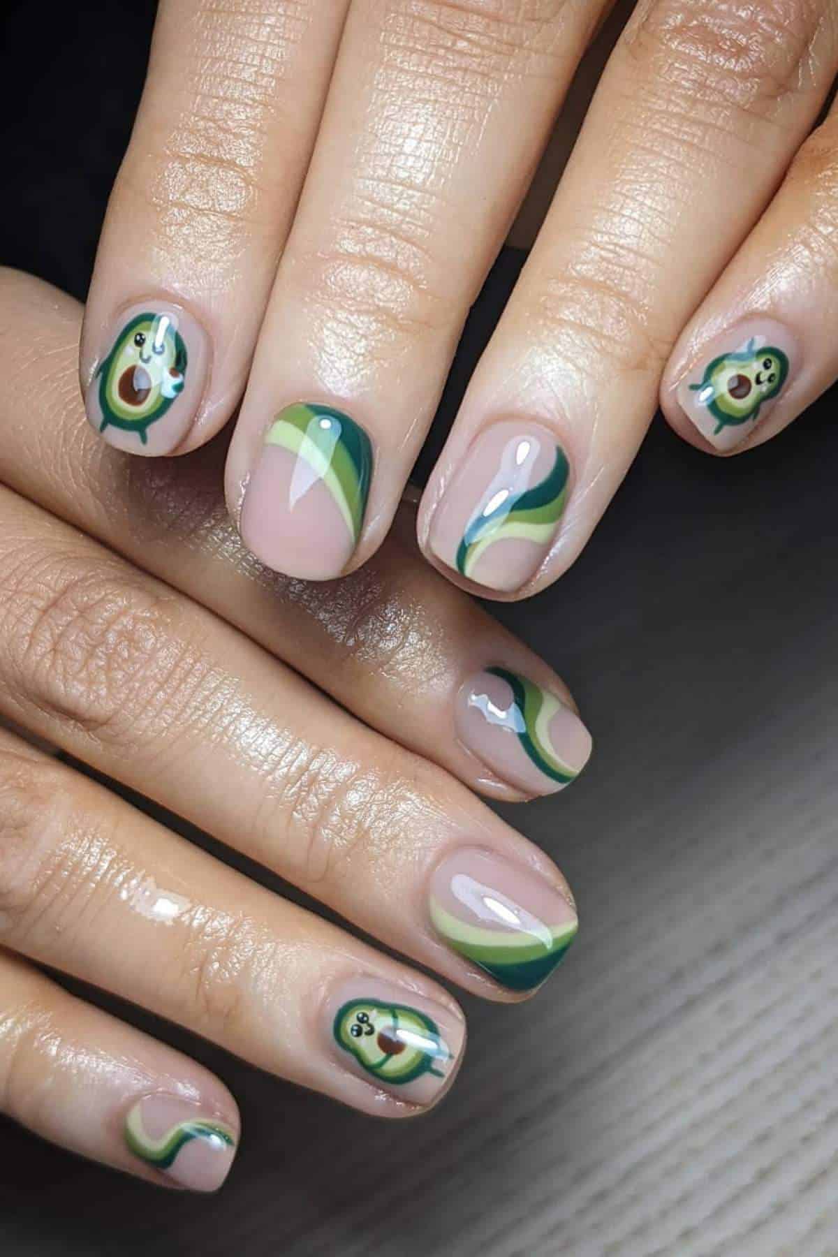 Close-up of hands with manicured nails featuring avocado designs and green wavy patterns on a nude base.