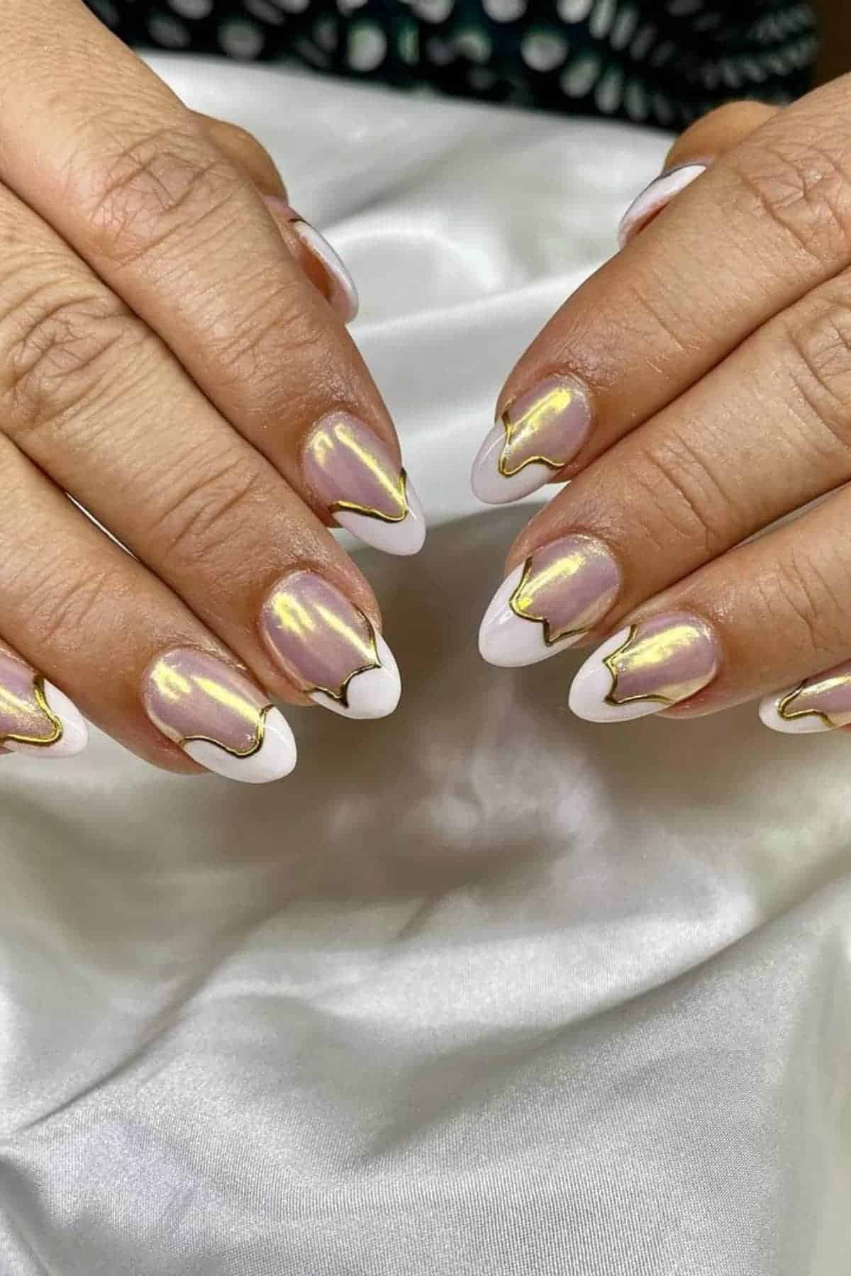 Hands with almond-shaped nails featuring a nude base, gold chrome accents, and white tips, resting on white fabric.