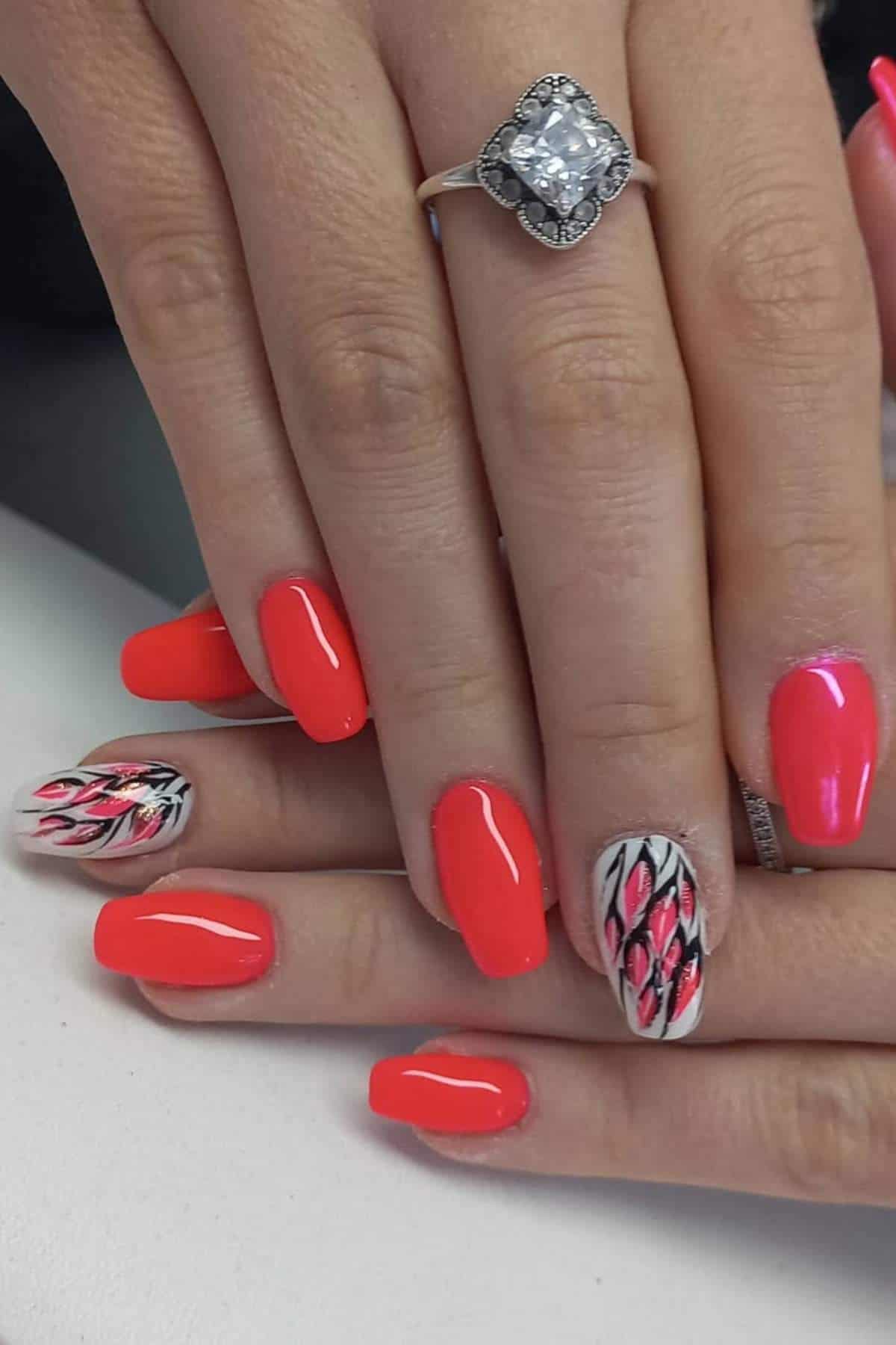 Hands with bright red manicured nails, two accent nails featuring black and pink flame-like designs, and a silver ring with a large square gemstone on one finger.