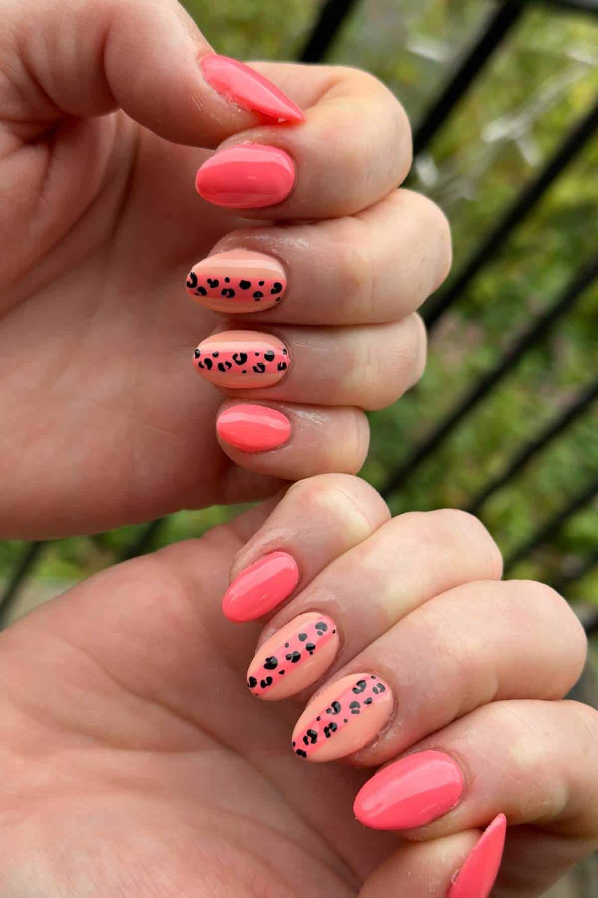 Hands with almond-shaped nails painted coral pink; some nails have black leopard print accent designs. Fence and greenery in the blurred background.