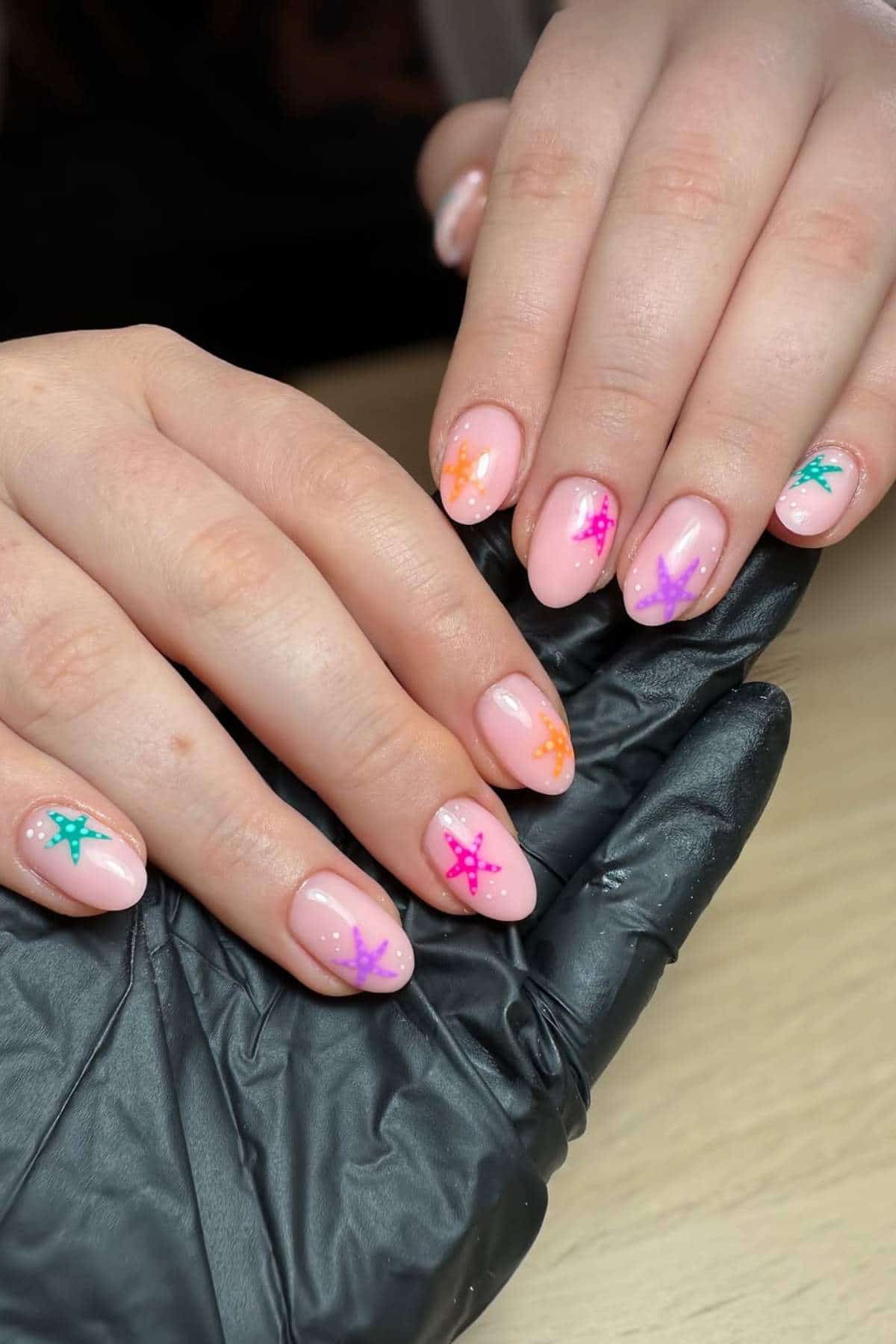 Hands with oval-shaped nails painted light pink, each nail decorated with colorful star designs in green, orange, pink, and purple. One hand rests on a black gloved hand.