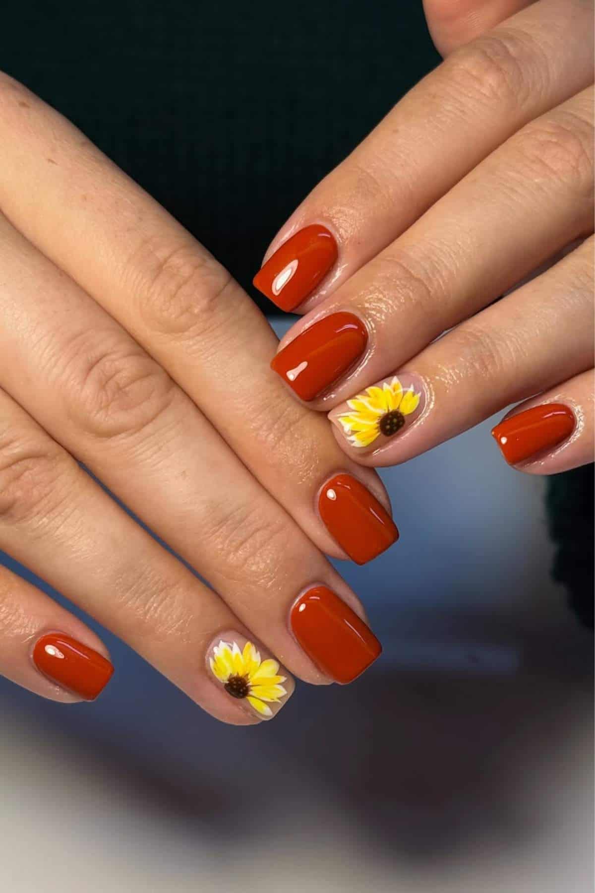 Hands with short, square-shaped nails painted in glossy burnt orange, with accent nails featuring yellow sunflower designs on a neutral base.