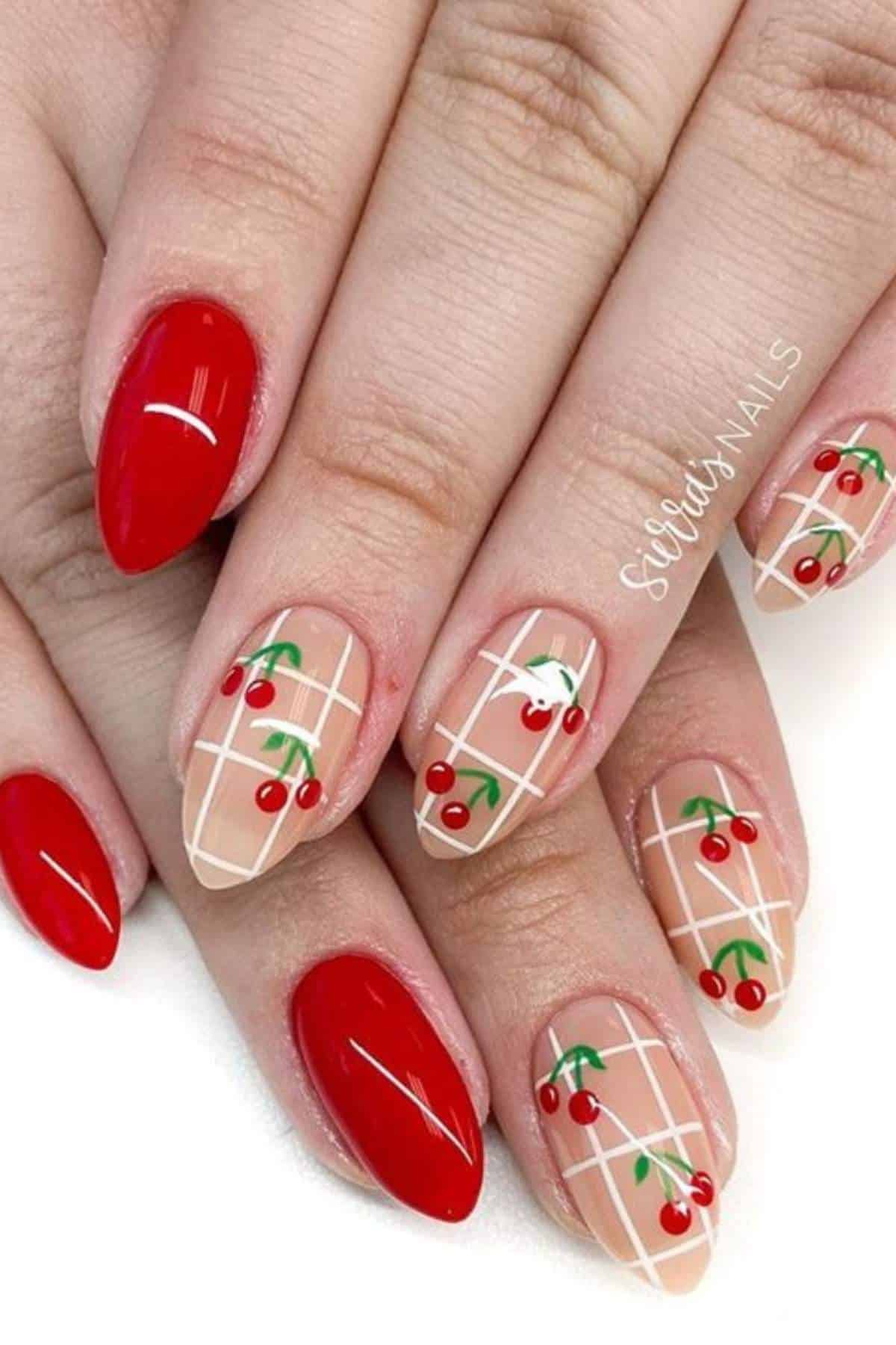 Hands with almond-shaped nails; some nails are painted red, others have a nude base with white grid lines and red cherry designs with green stems.
