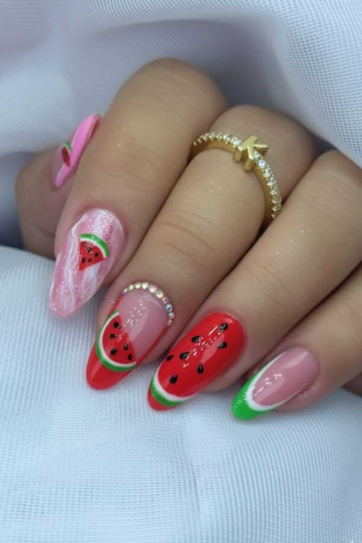 A hand with long nails painted in a watermelon theme, featuring red, green, and pink colors, rhinestone accents, and a gold ring with the letter "K.