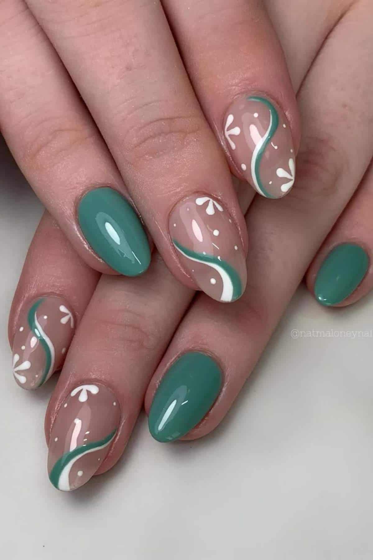 Close-up of hands with almond-shaped nails; some painted solid green, others with green and white abstract swirls and small white flower designs on a nude base.