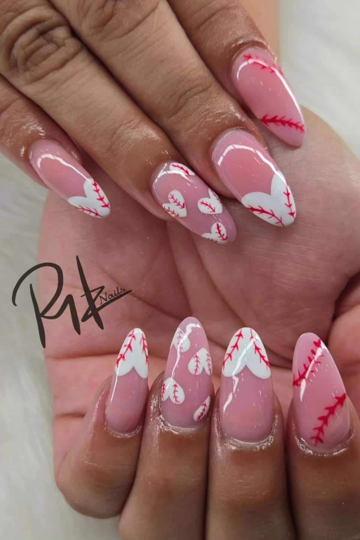 Hands with long, almond-shaped nails featuring pink and white baseball-themed nail art with red stitching details.