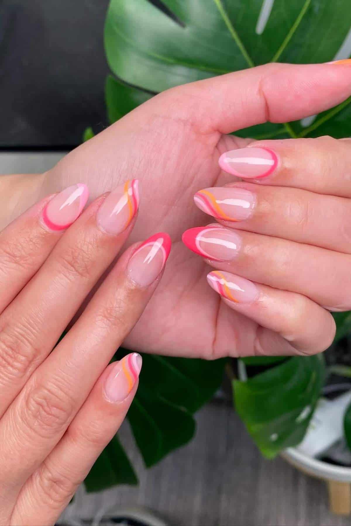 Hands with almond-shaped nails featuring pink, orange, and yellow wavy designs on a clear base, shown against a background with green leaves.