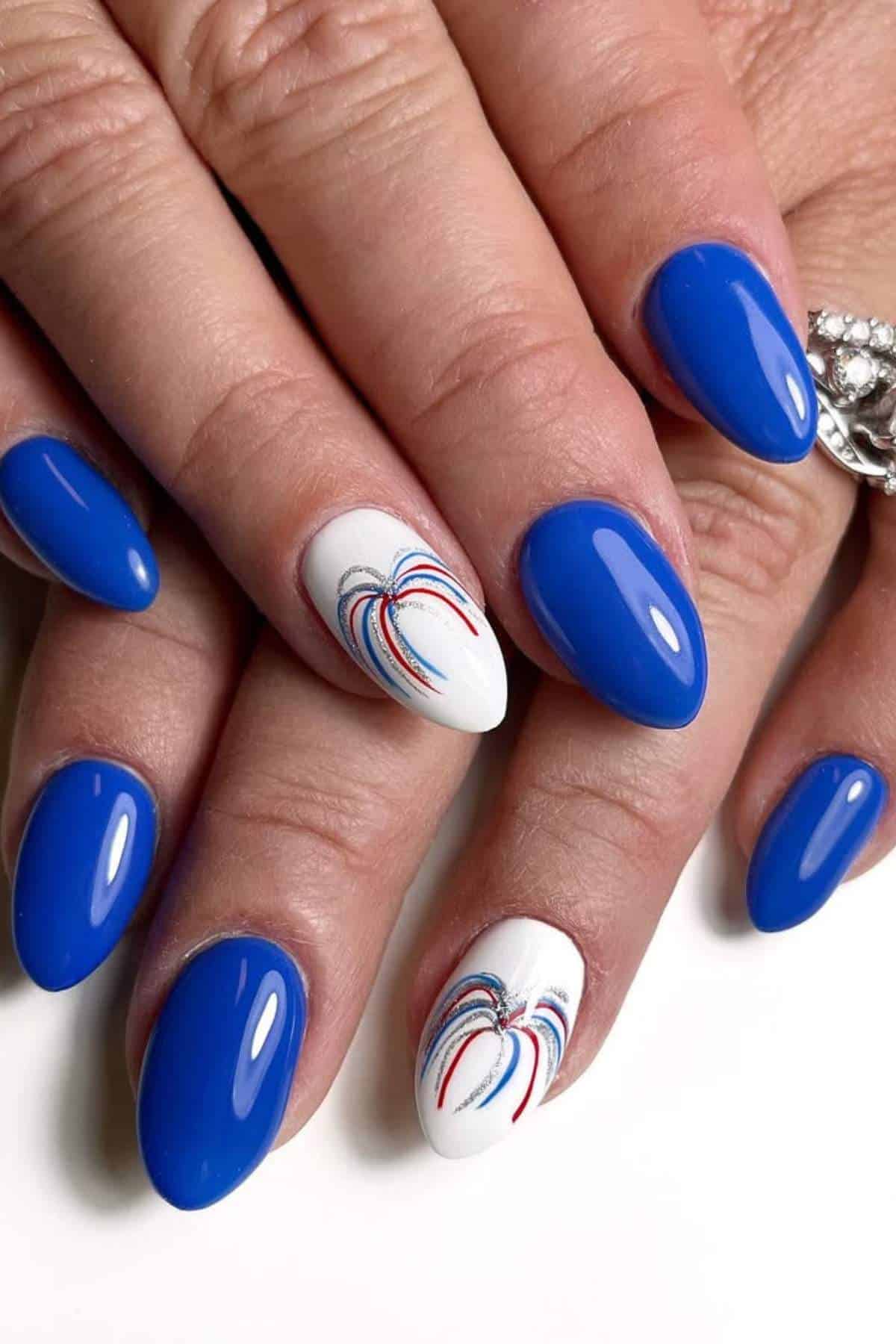 Oval nails with a glossy royal blue polish, featuring two accent nails with white polish and red, white, and blue firework designs.