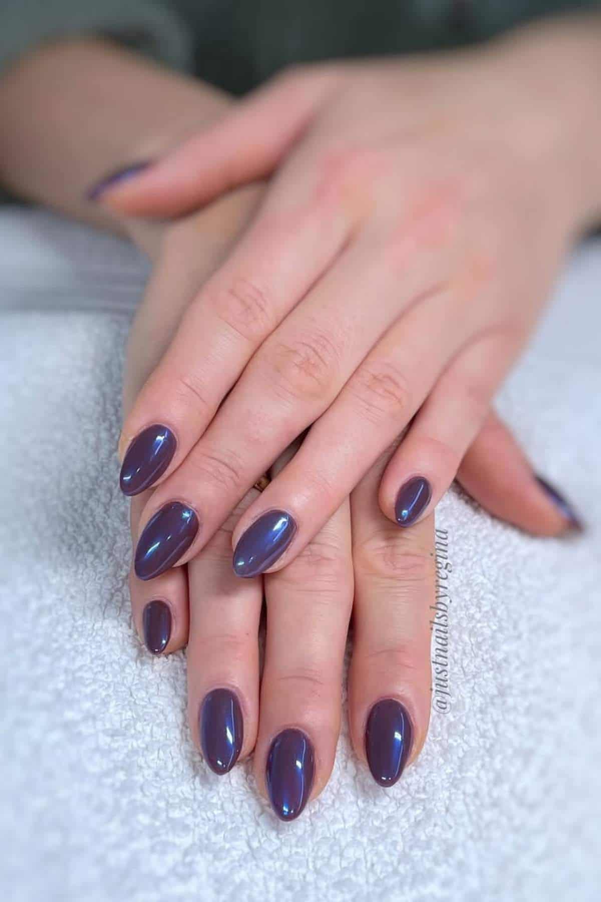 Close-up of hands with oval-shaped nails painted in a shiny, metallic purple polish, resting on a white textured surface.